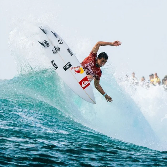 A surfer in a red patterned shirt rides a white surfboard with multiple sponsor logos, including Quiksilver and Red Bull, over a breaking turquoise wave with spectators visible in the background.