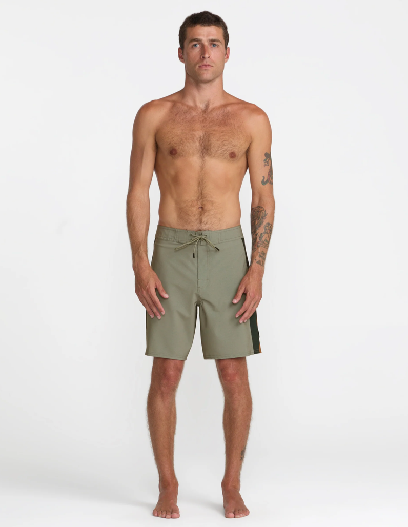 A male model stands barefoot against a plain light gray studio background wearing olive green board shorts with a drawstring waist and a dark side stripe.