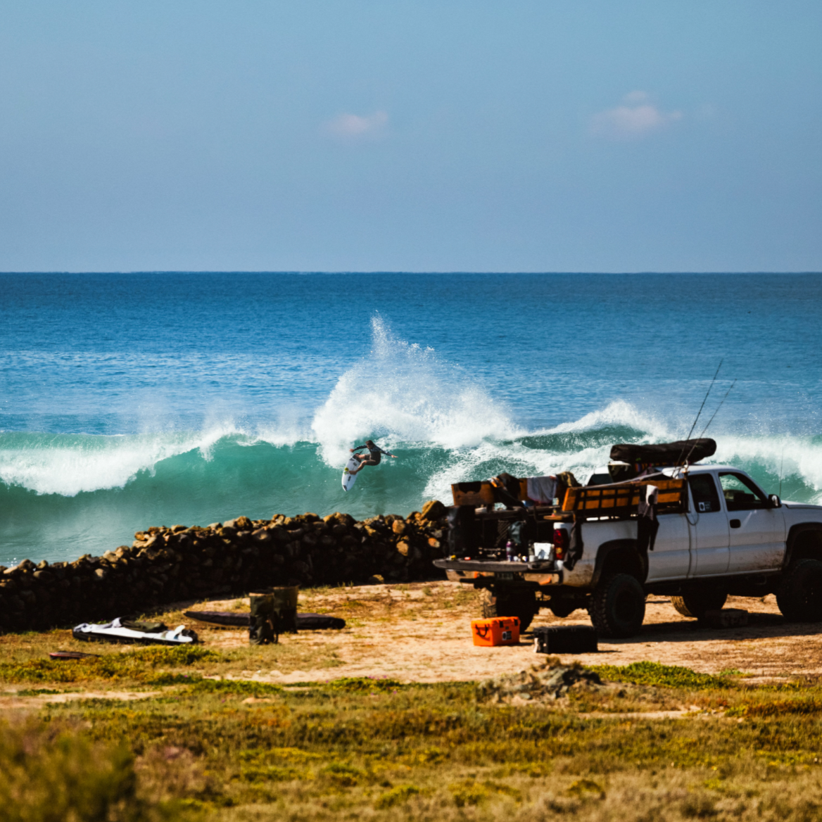 A surfer rides a breaking ocean wave while a white pickup truck loaded with gear sits on a sandy beachside spot. Rocks line the shore, and the blue ocean and sky fill the background.
