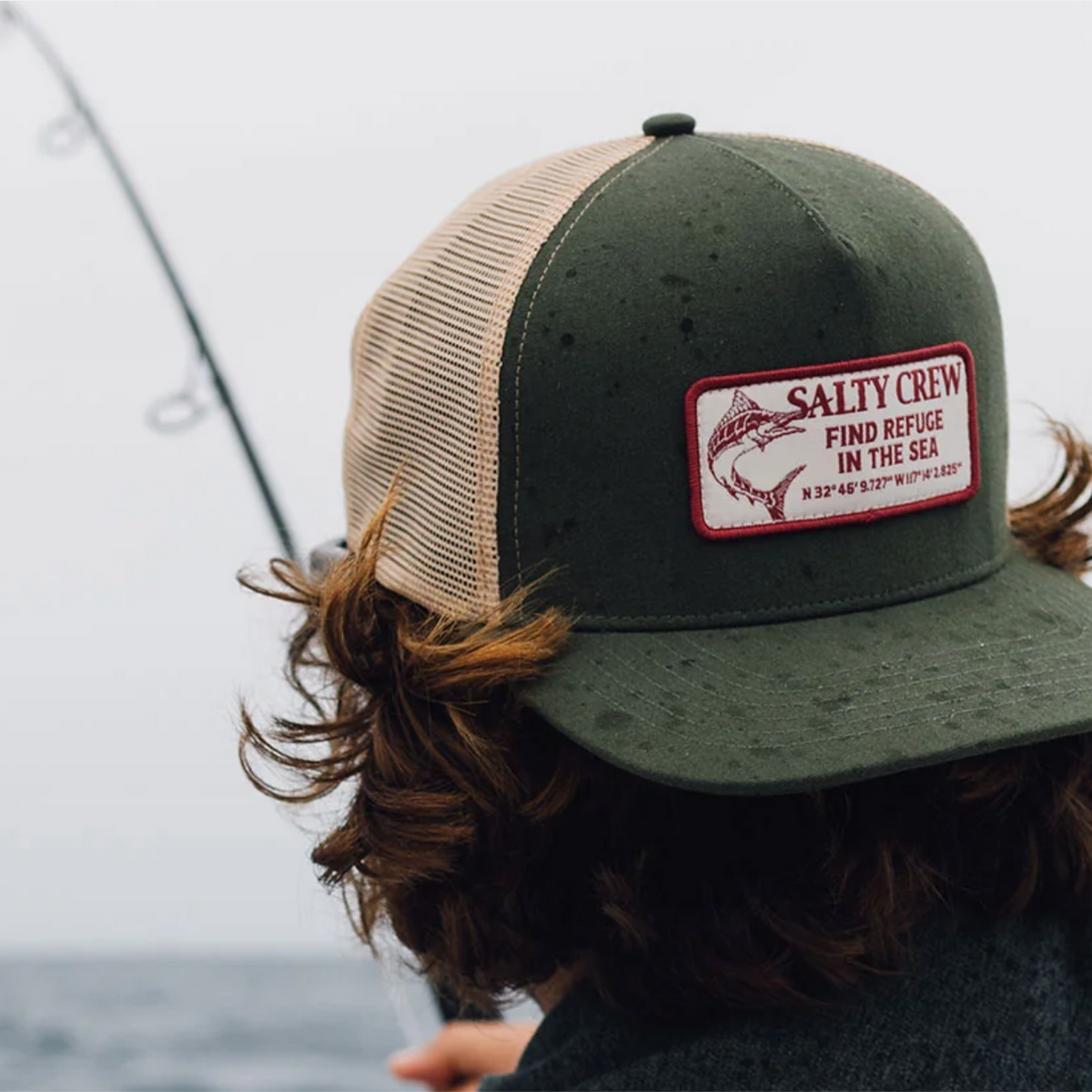 A person wearing an olive green and beige trucker hat with a 'Salty Crew' patch, photographed from behind with a fishing rod and overcast sky in the background.