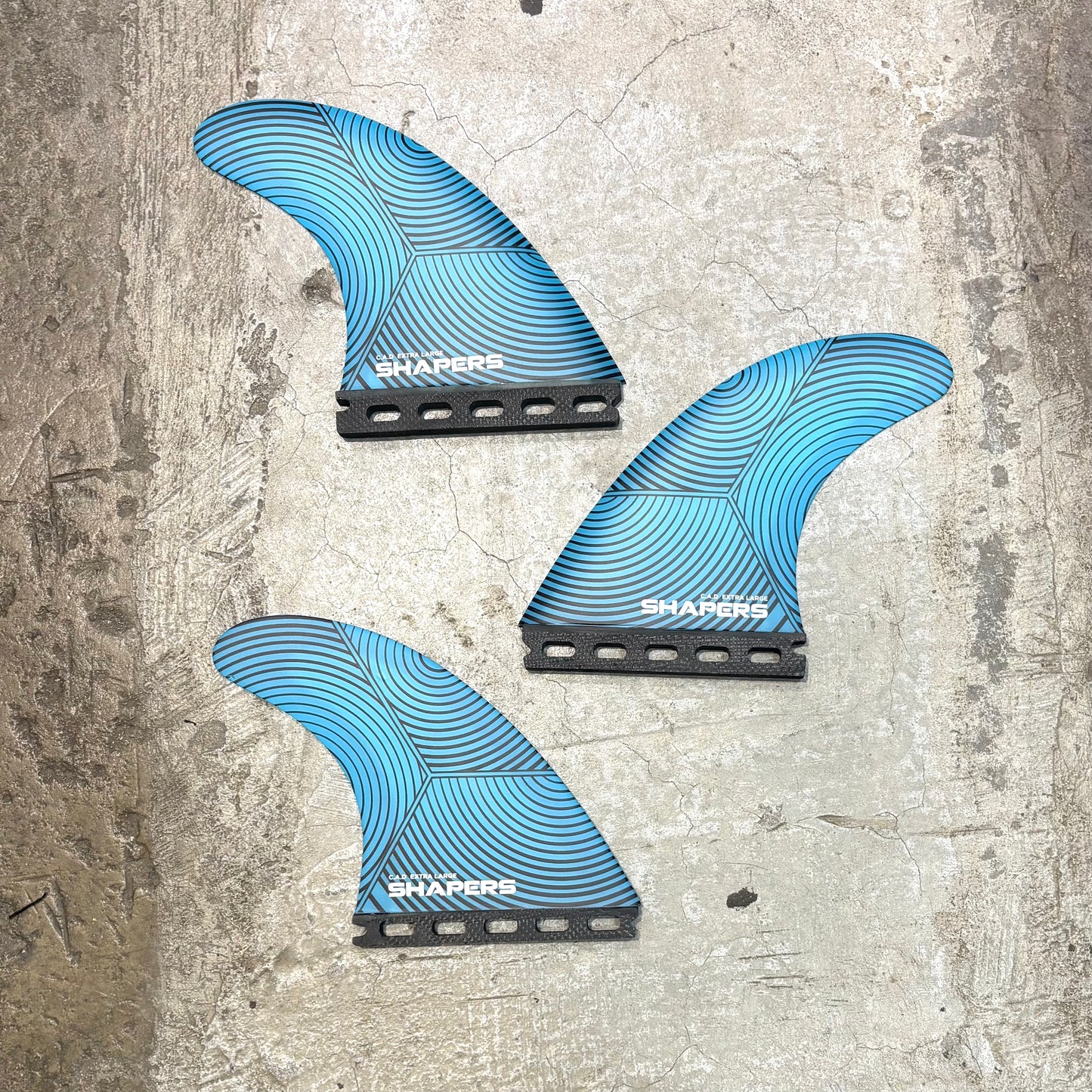Three blue surfboard fins with 'Shapers' branding on a textured gray surface.