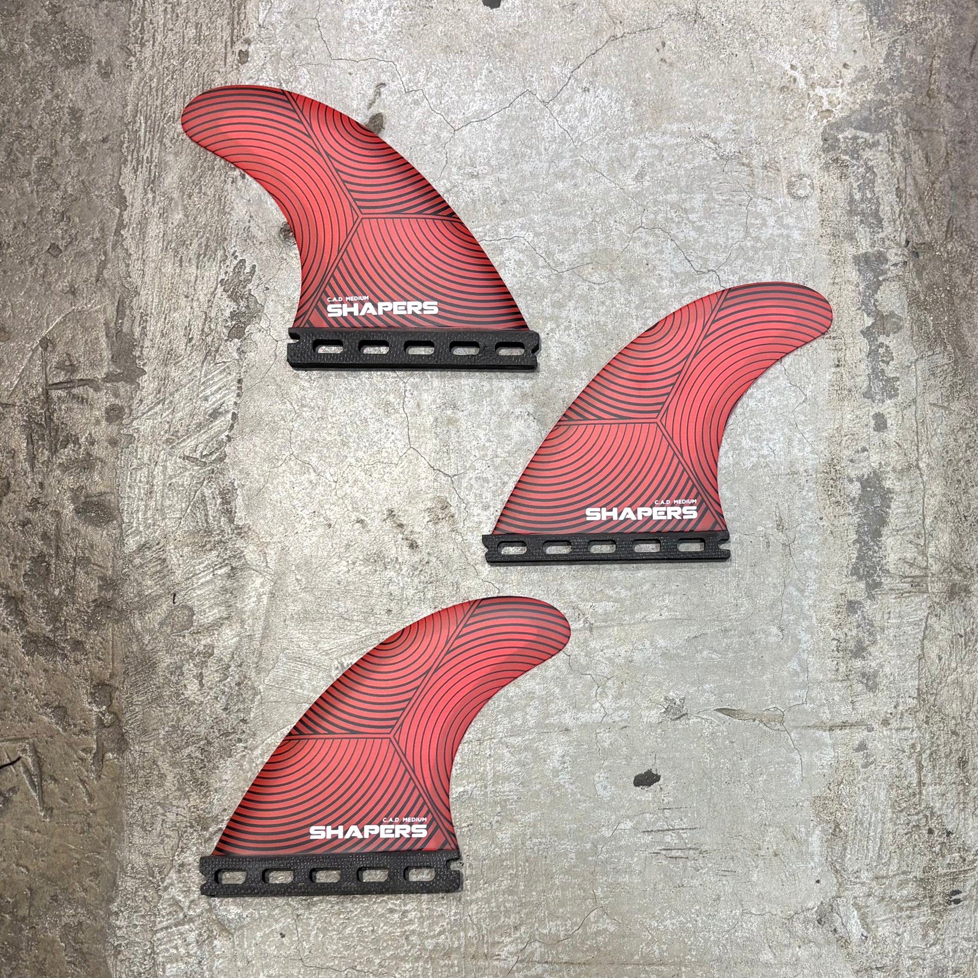 Three red surfboard fins with 'Shapers' branding on a textured gray surface