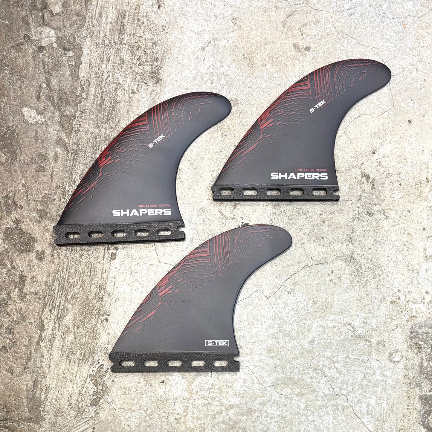 Three surfboard fins with red patterns on a textured surface, branded 'Shapers'.