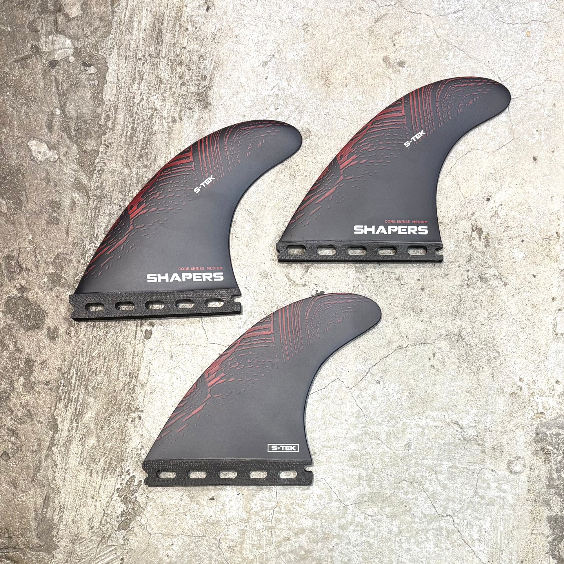 Three surfboard fins with red patterns on a textured surface, branded 'Shapers'.