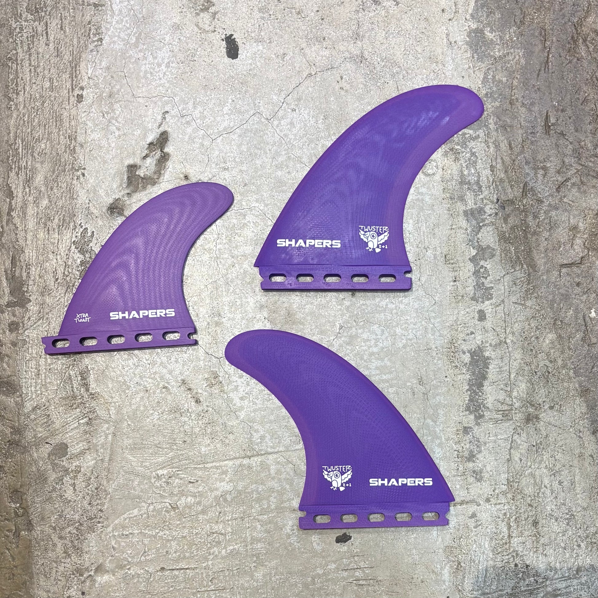Three purple surfboard fins with 'Shapers' branding on a textured surface.