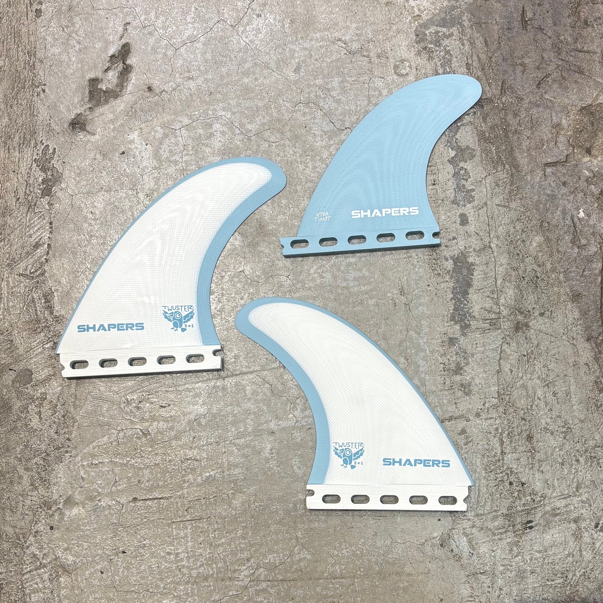 Three surfboard fins with 'Shapers' branding on a textured surface
