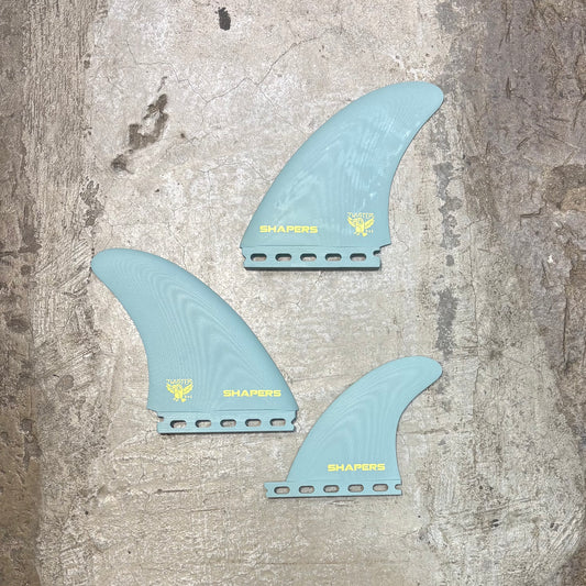 Three blue surfboard fins with 'Shapers' branding on a textured surface