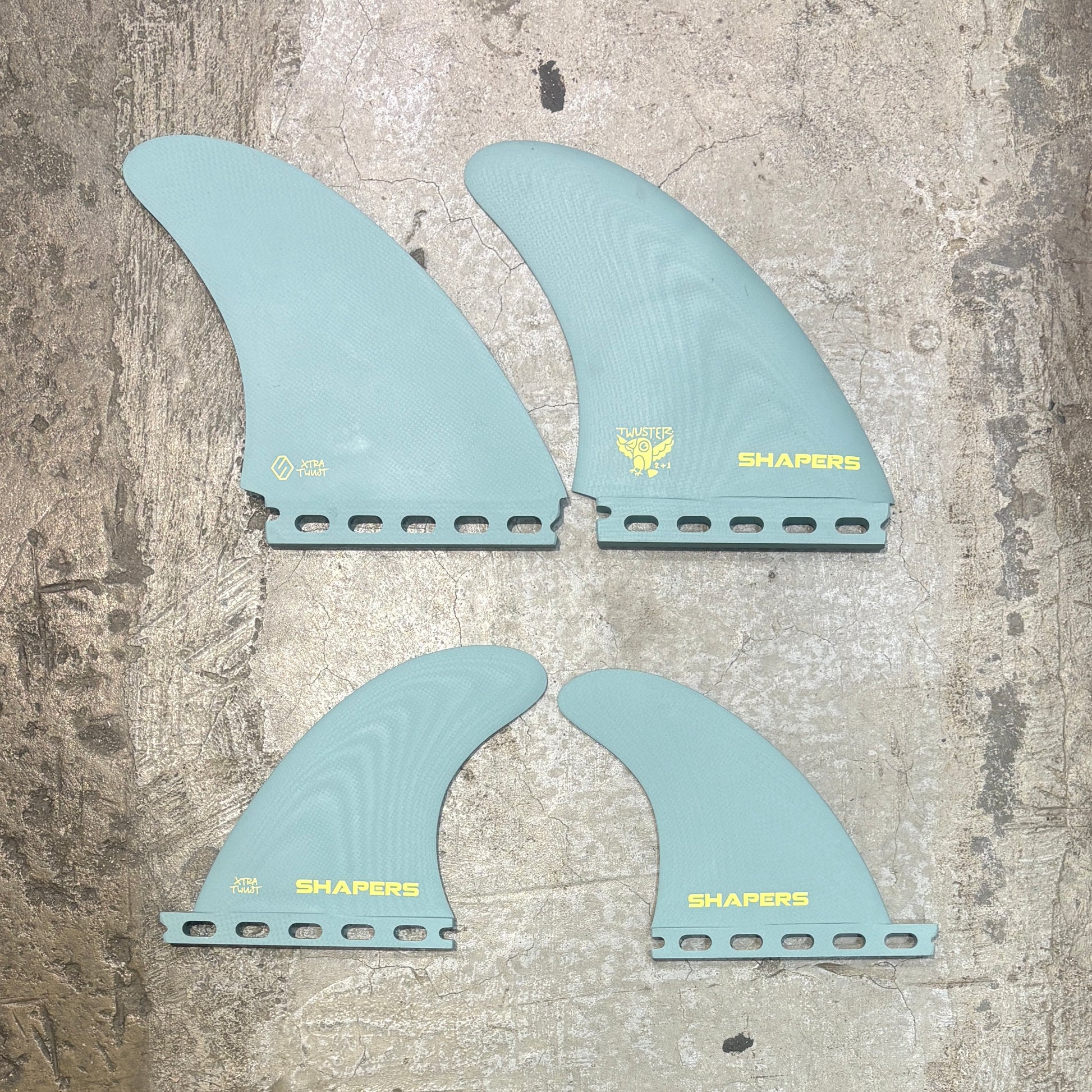 Four light blue surfboard fins with 'SHAPERS' branding on a textured surface.