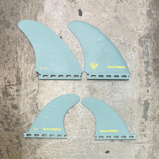 Four light blue surfboard fins with 'SHAPERS' branding on a textured surface.