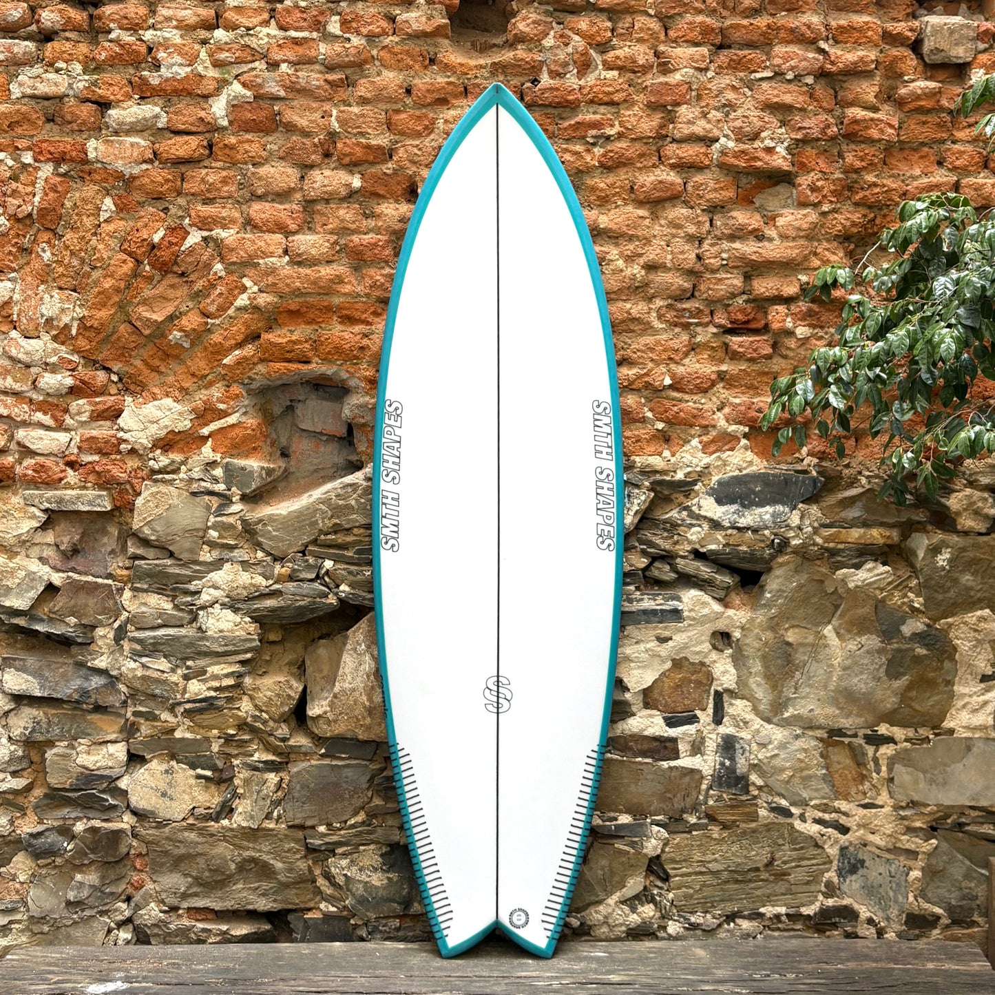 White SMTH surfboard with blue edges against a rustic stone wall.