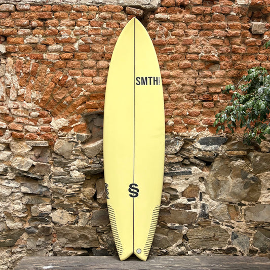 Yellow surfboard with 'SMTH' branding against a rustic stone wall.