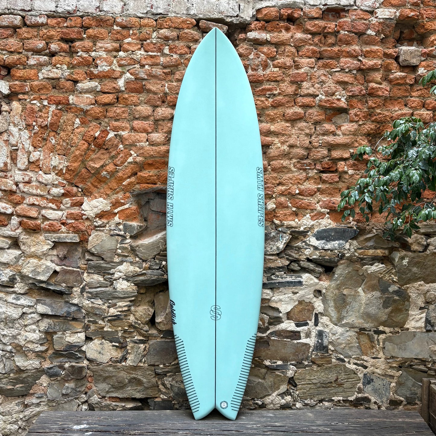 Light blue SMTH surfboard leaning against a rustic stone wall.