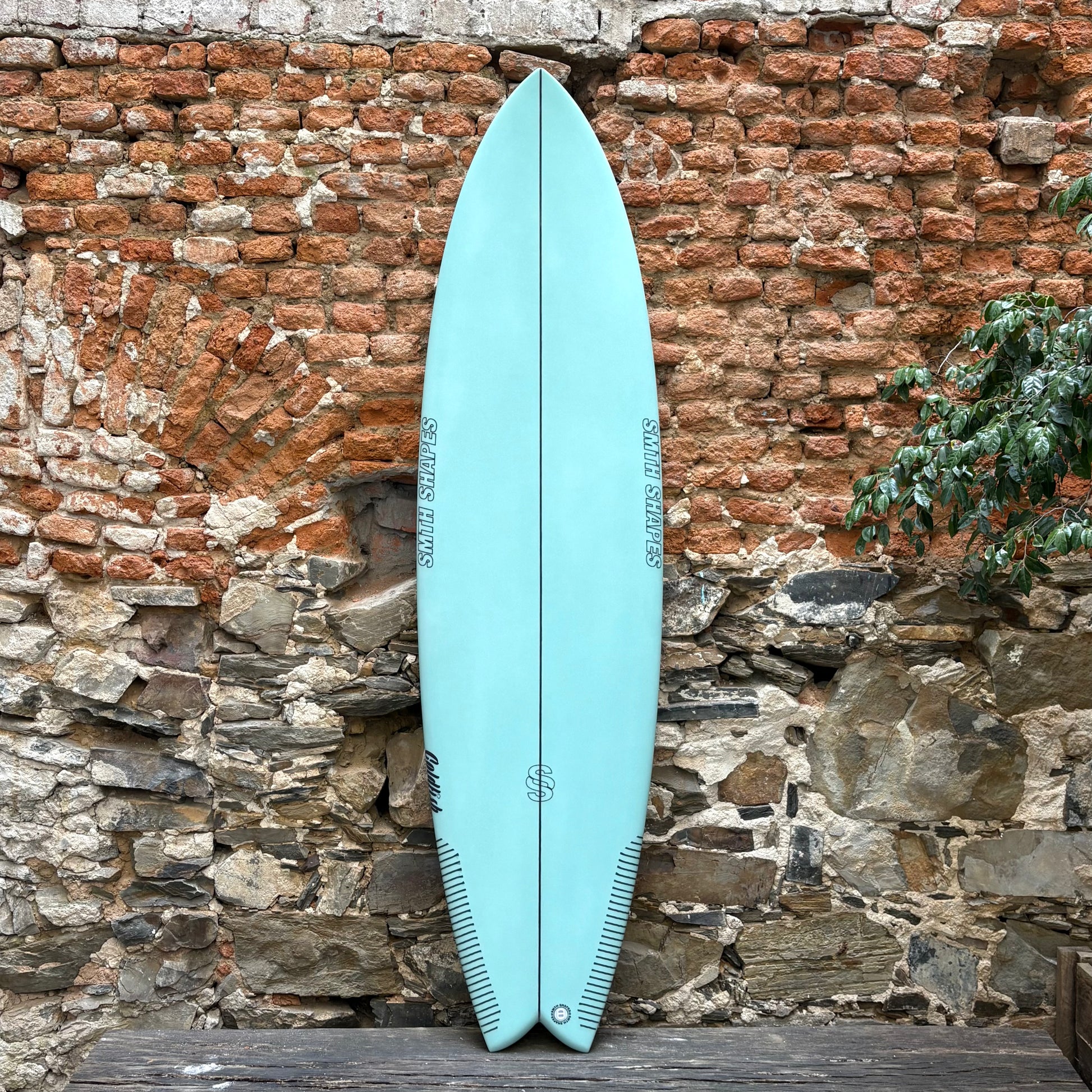 Light blue SMTH surfboard leaning against a rustic stone wall.