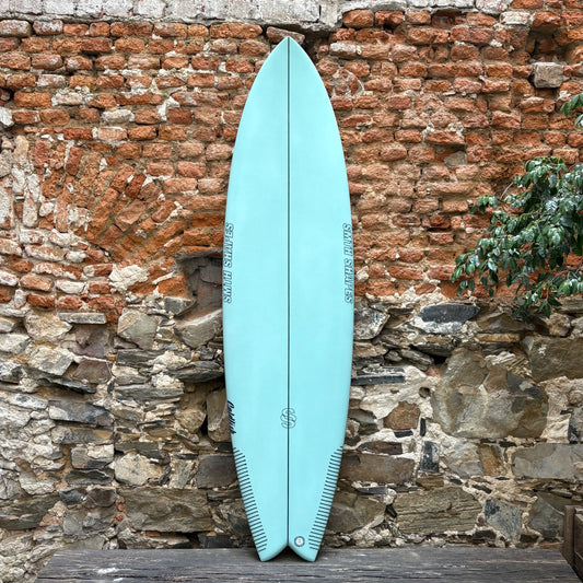 Light blue SMTH surfboard leaning against a rustic stone wall.