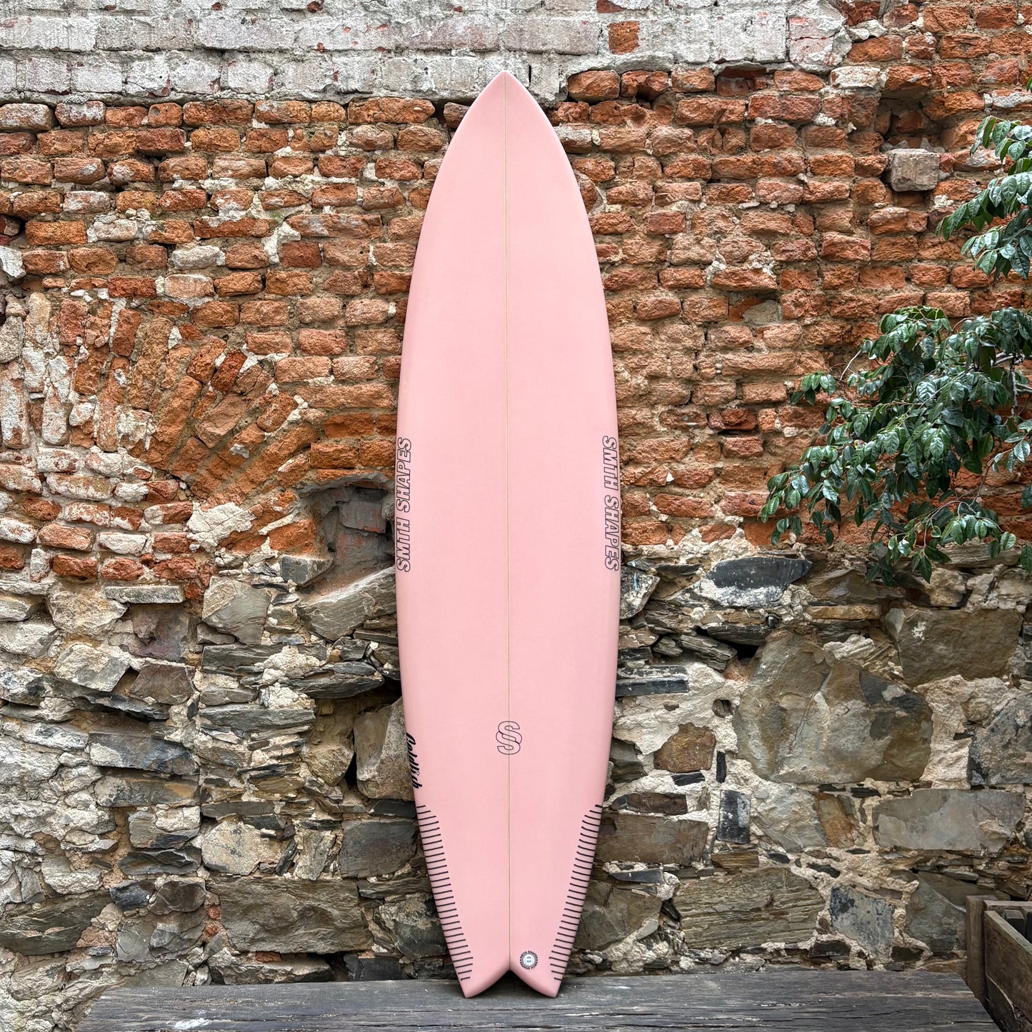 Pink SMTH surfboard leaning against a rustic stone wall with a plant in the background