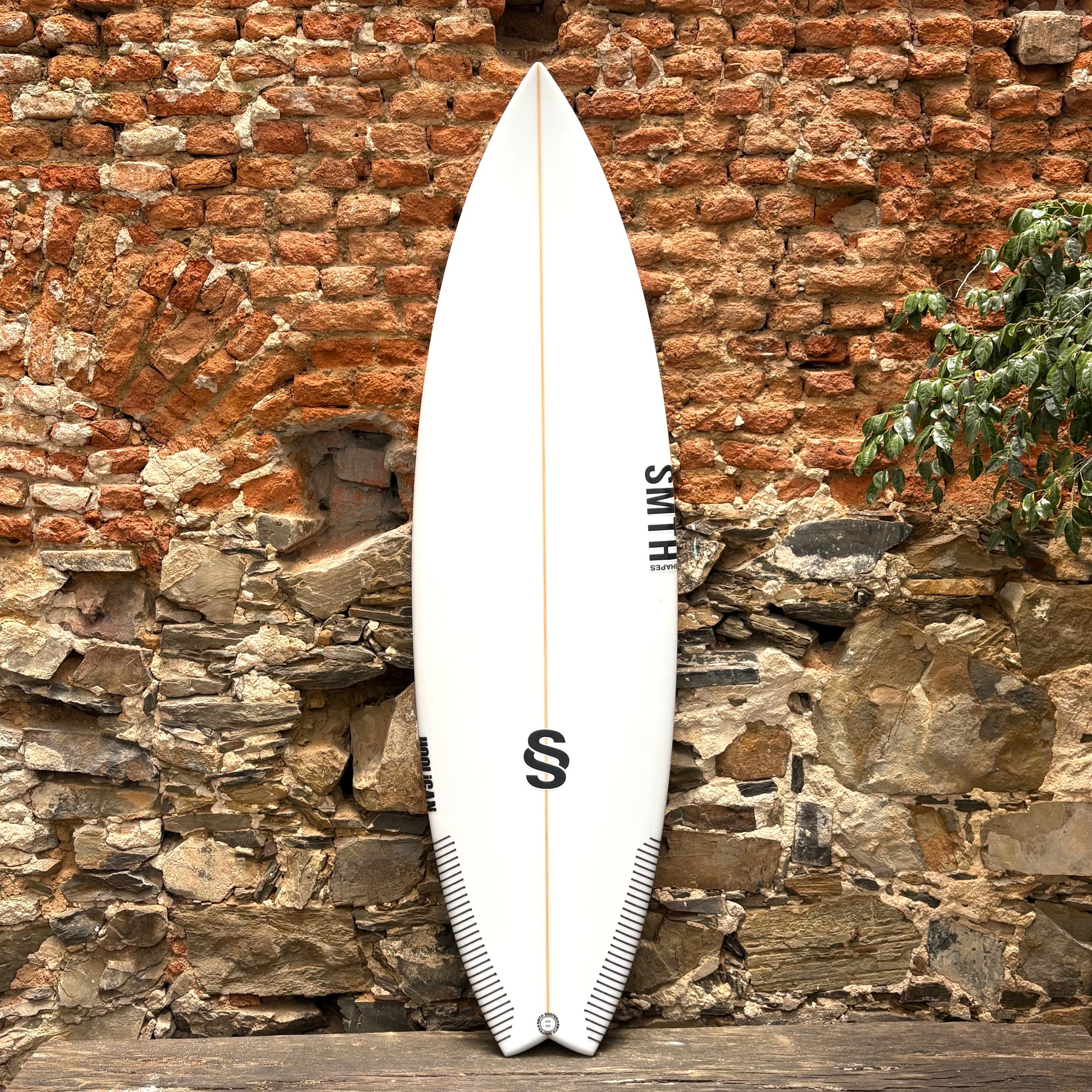 White Hooligan surfboard with 'Smth' branding against a rustic stone wall.