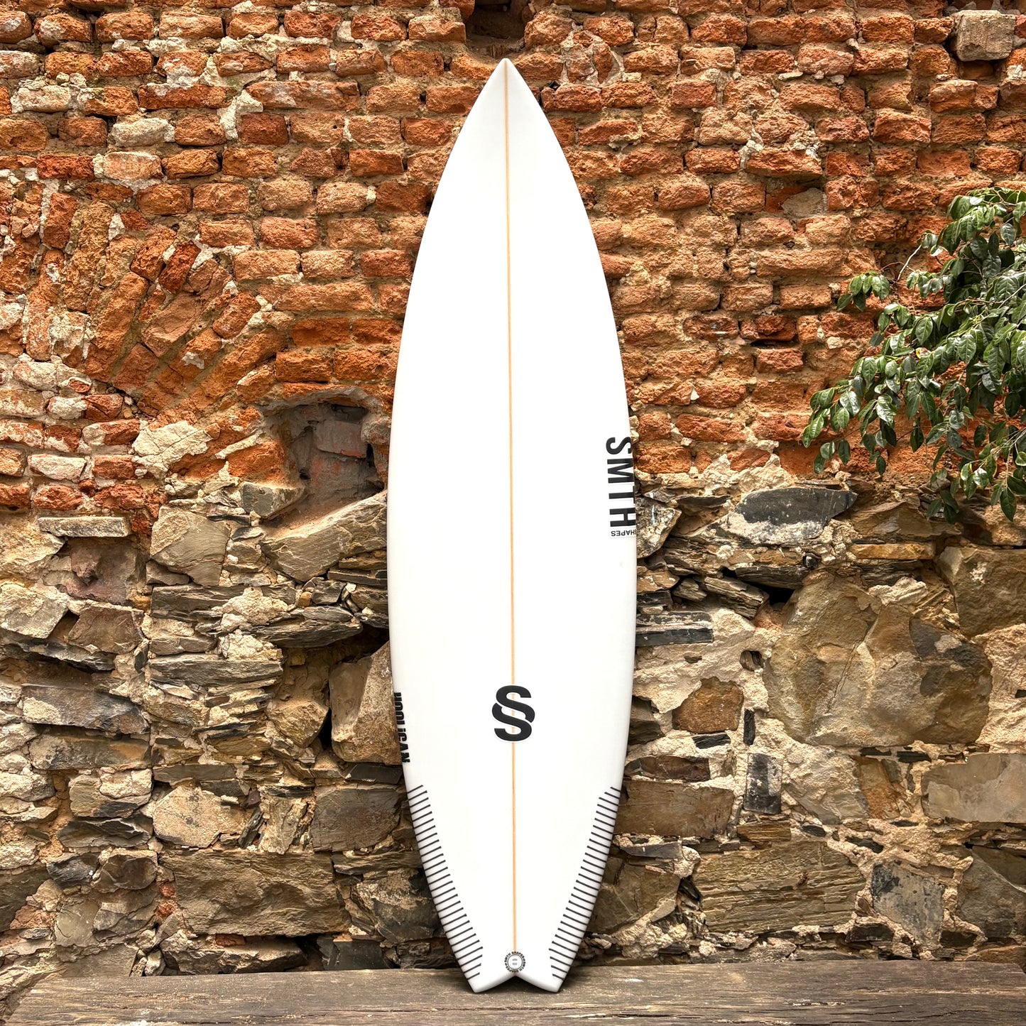 White SMTH surfboard in front of rustic stone wall.