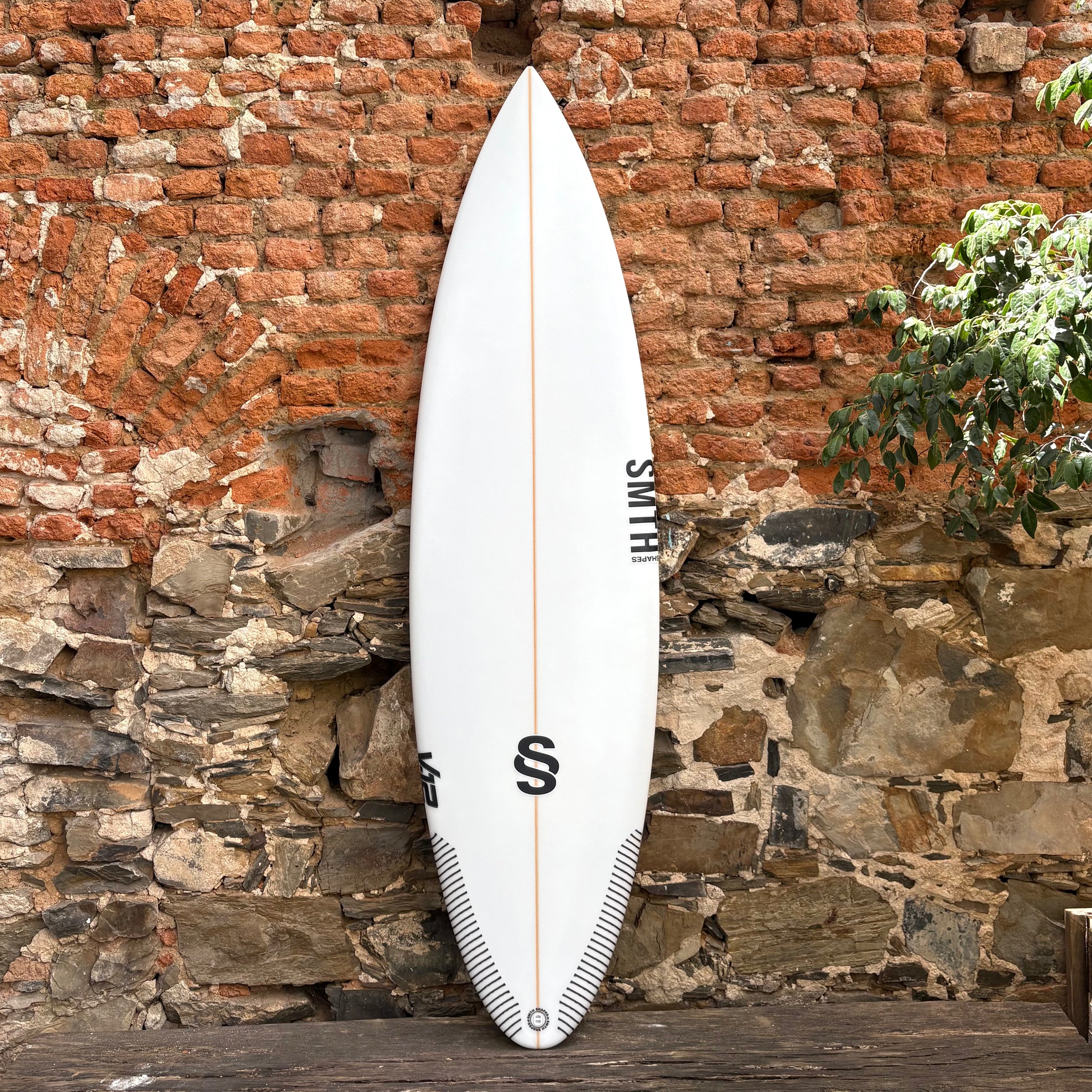 White SMTH V12 surfboard with a black logo against a rustic stone wall.