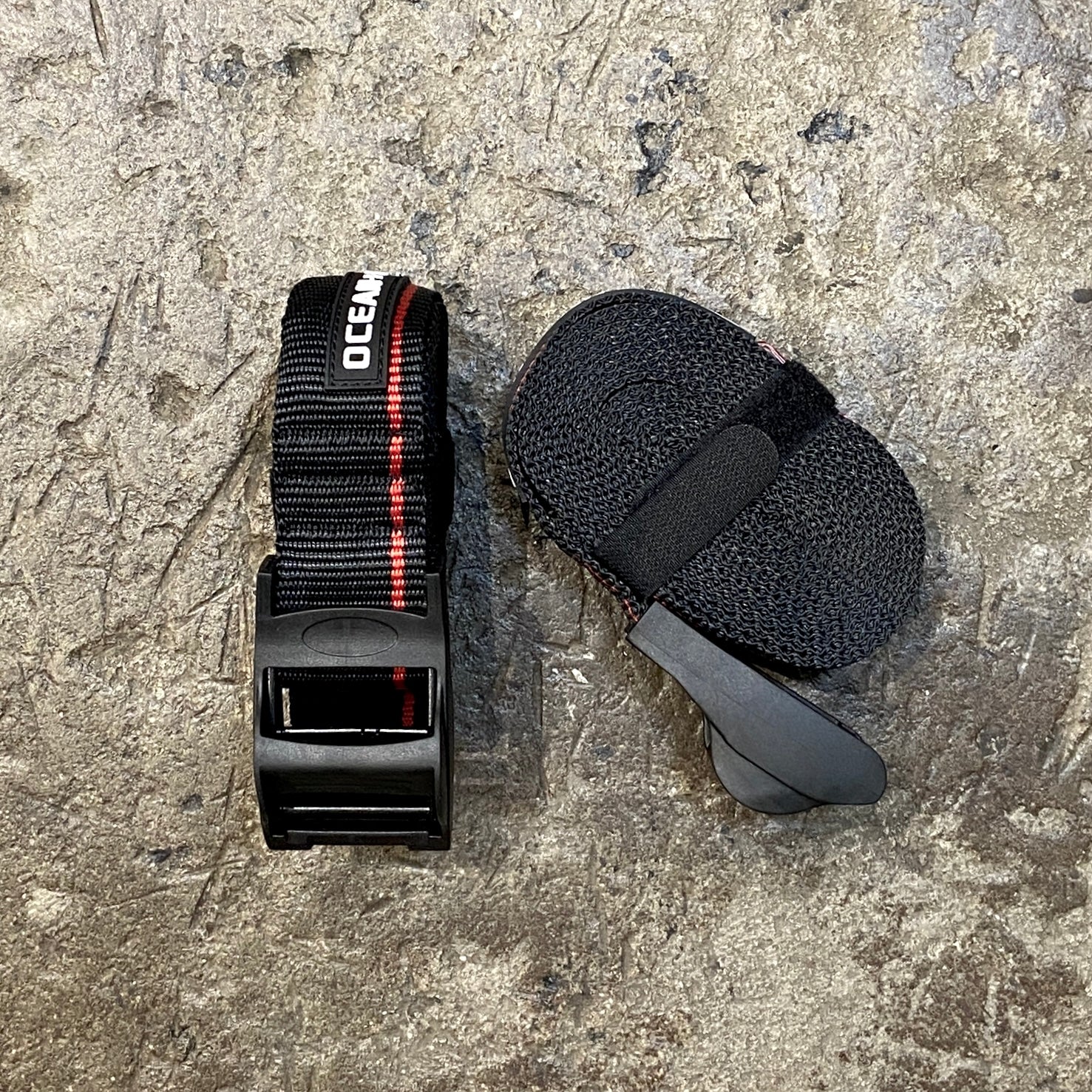 A black and red surfboard tie down strap with a protective rubber cam buckle and Velcro loop, placed on a textured grey surface.