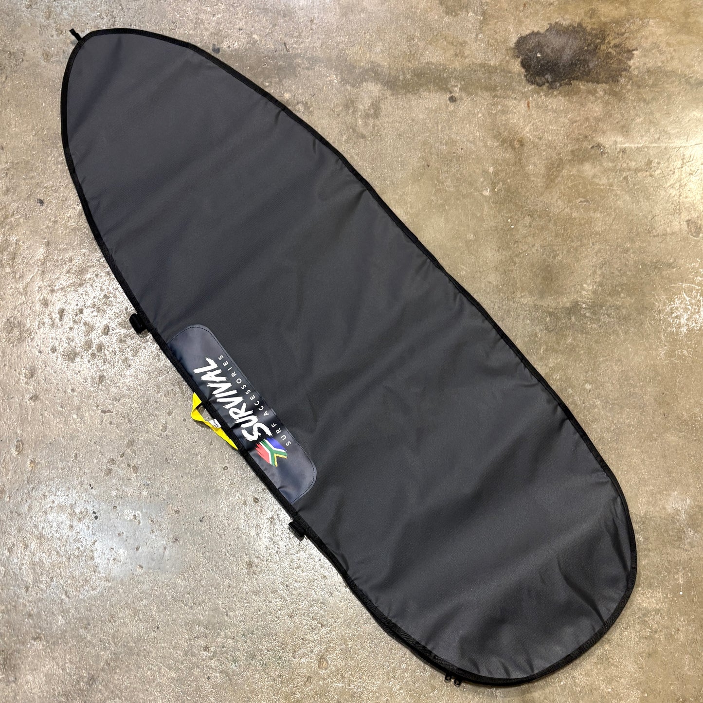 A black surfboard carrying bag laid flat on a concrete floor, with a visible SURVIVAL brand patch near the lower-middle section.