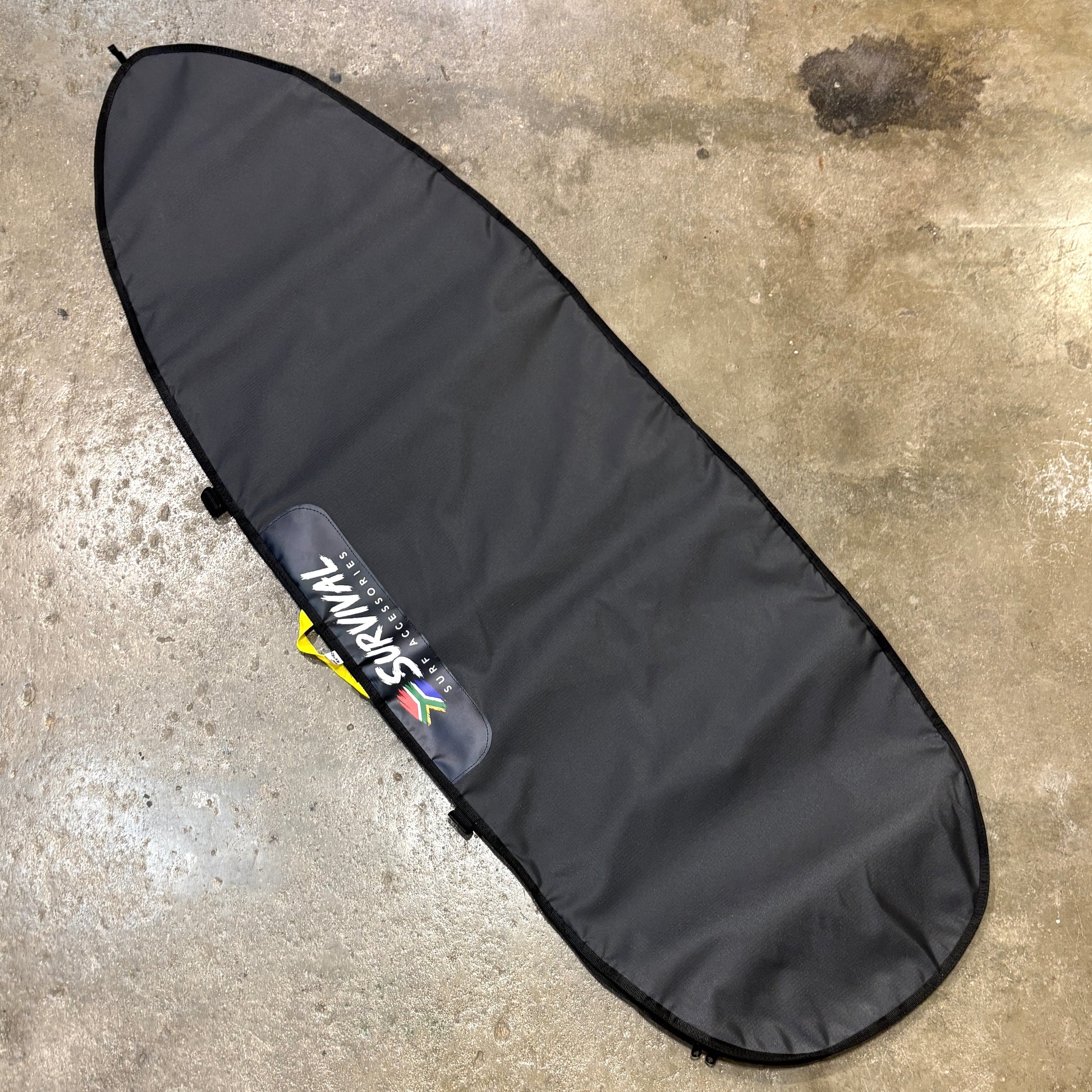A black surfboard carrying bag laid flat on a concrete floor, with a visible SURVIVAL brand patch near the lower-middle section.