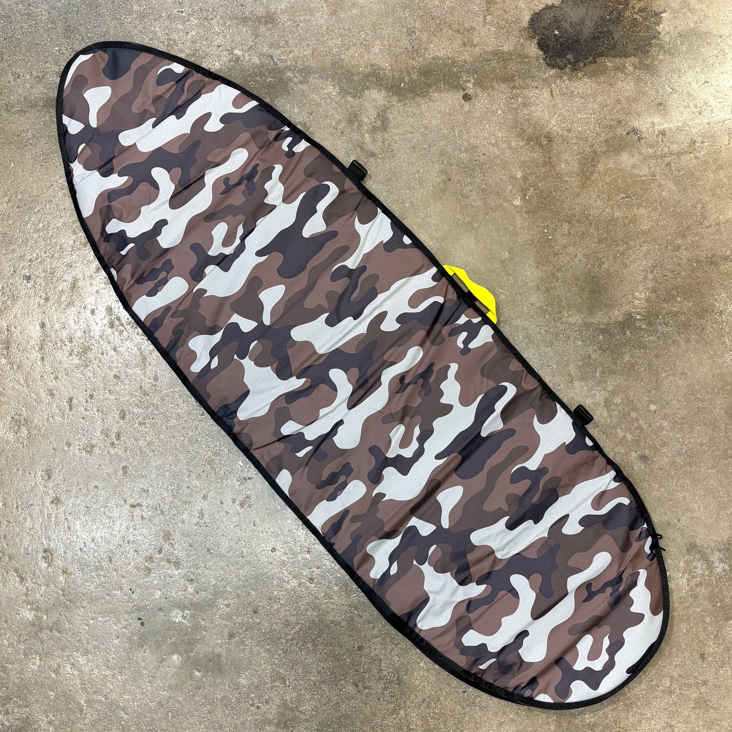 A surfboard carrying case or padded board cover with a brown, gray, and white camouflage pattern, laid diagonally on a concrete floor.