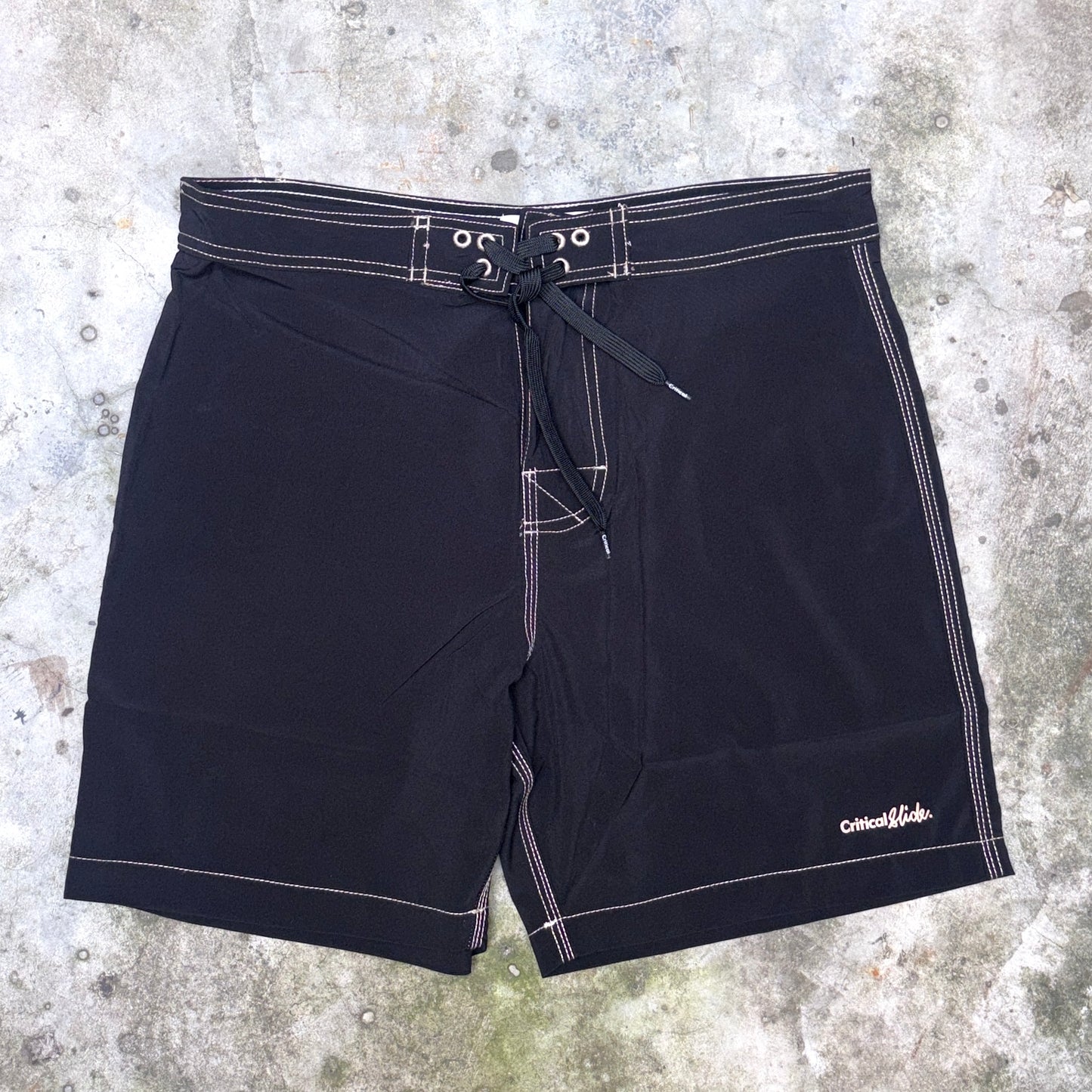 Black board shorts with white contrast stitching and a drawstring waist, laid flat on a textured concrete surface. The brand name 'Critical Velocity' is visible in white script on the lower right leg.