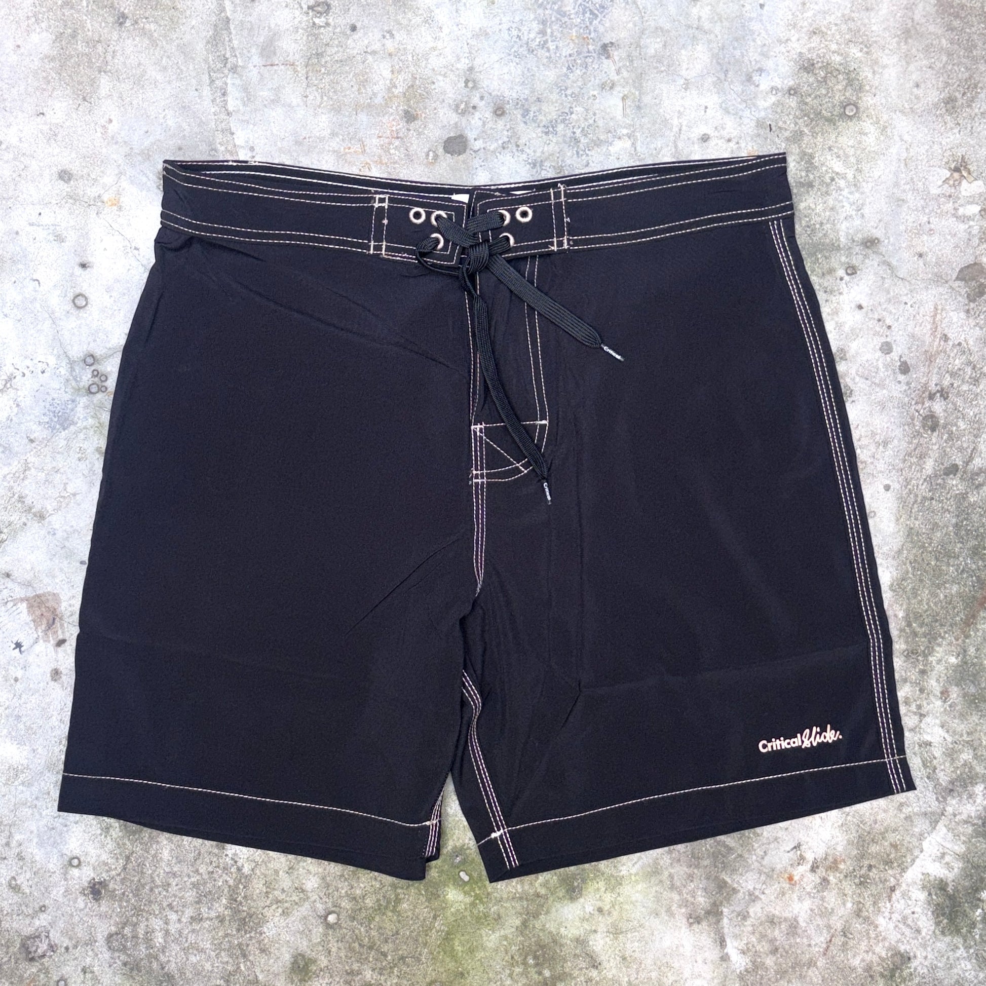 Black board shorts with white contrast stitching and a drawstring waist, laid flat on a textured concrete surface. The brand name 'Critical Velocity' is visible in white script on the lower right leg.