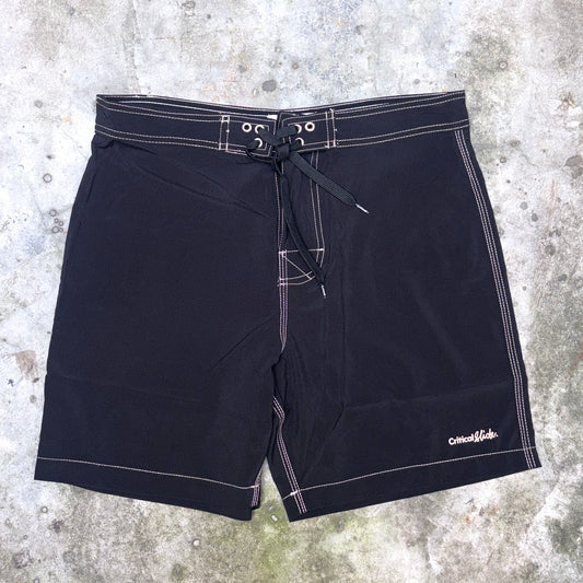 Black board shorts with white contrast stitching and a drawstring waist, laid flat on a textured concrete surface. The brand name 'Critical Velocity' is visible in white script on the lower right leg.