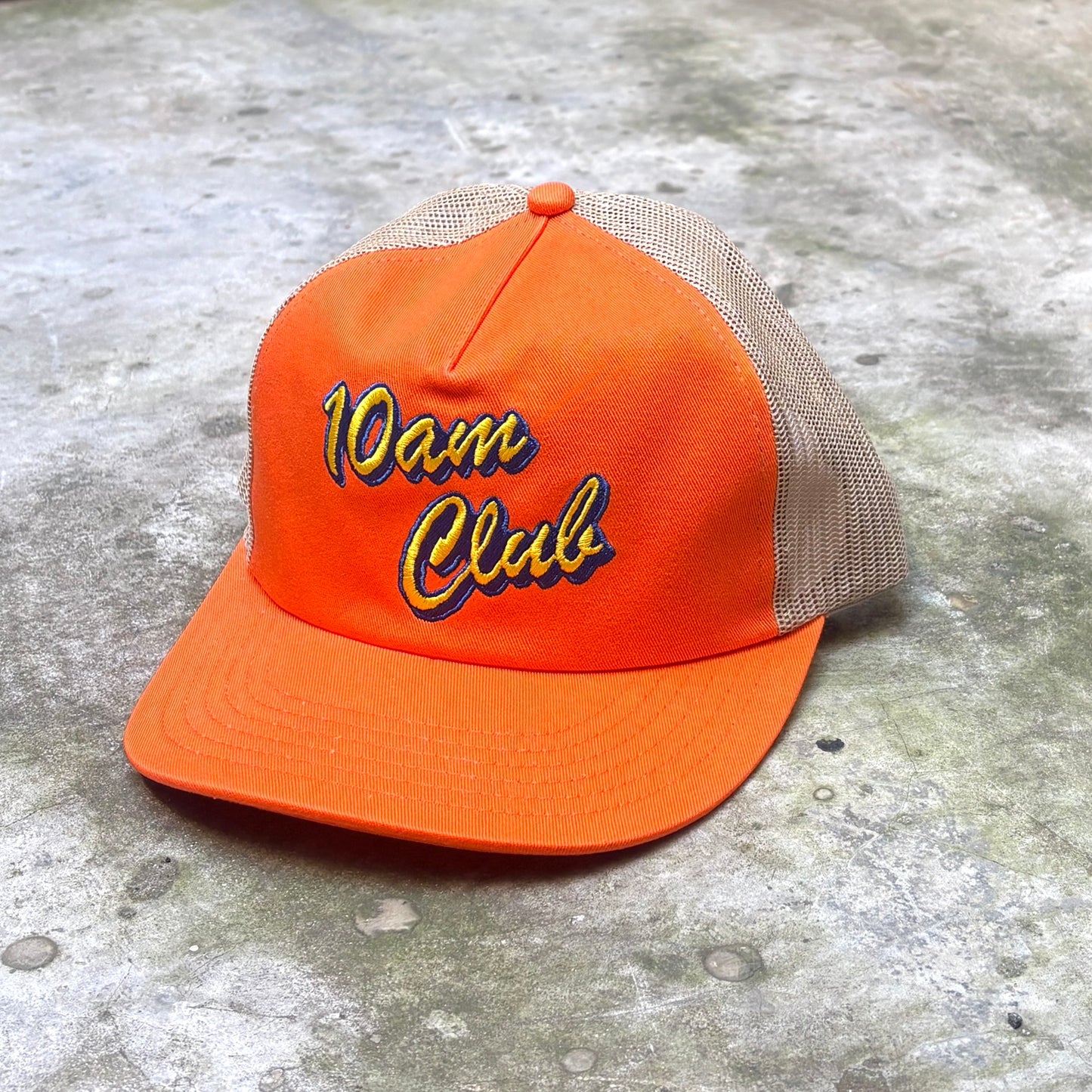 An orange trucker-style cap with beige mesh back panels and '10am Club' embroidered on the front in yellow and blue, placed on a textured gray concrete surface.