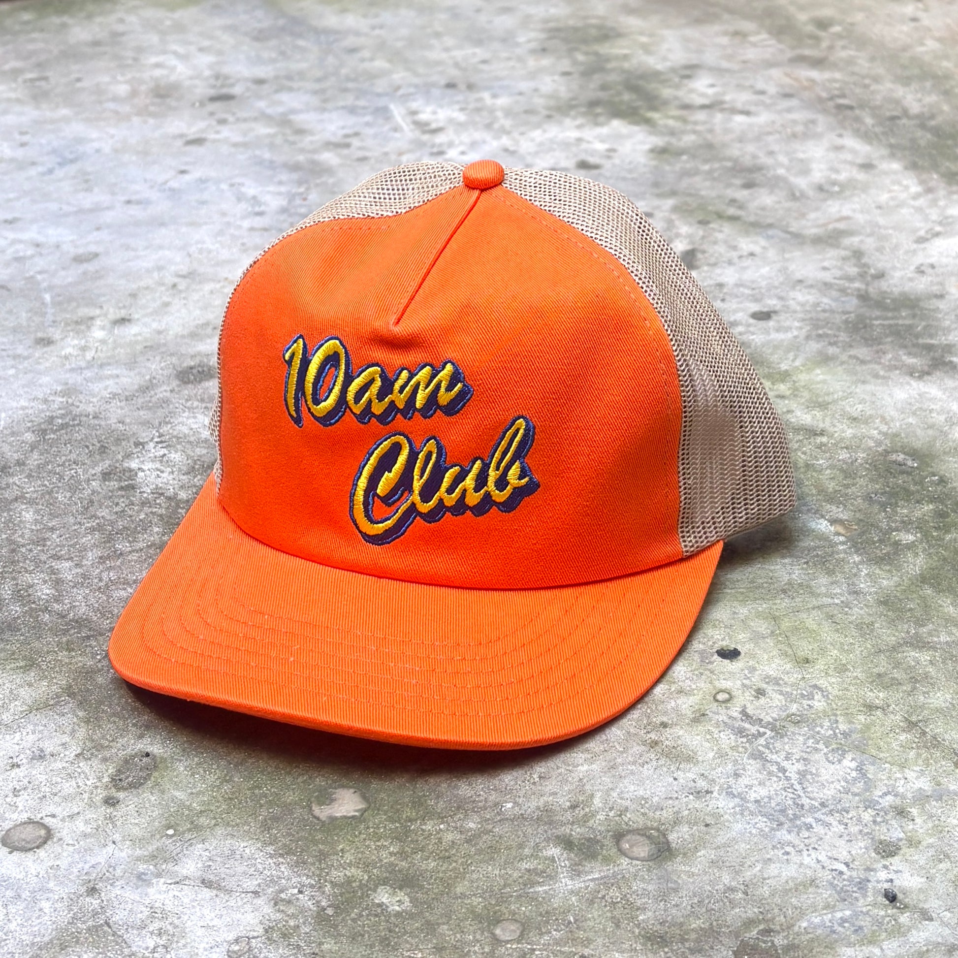 An orange trucker-style cap with beige mesh back panels and '10am Club' embroidered on the front in yellow and blue, placed on a textured gray concrete surface.