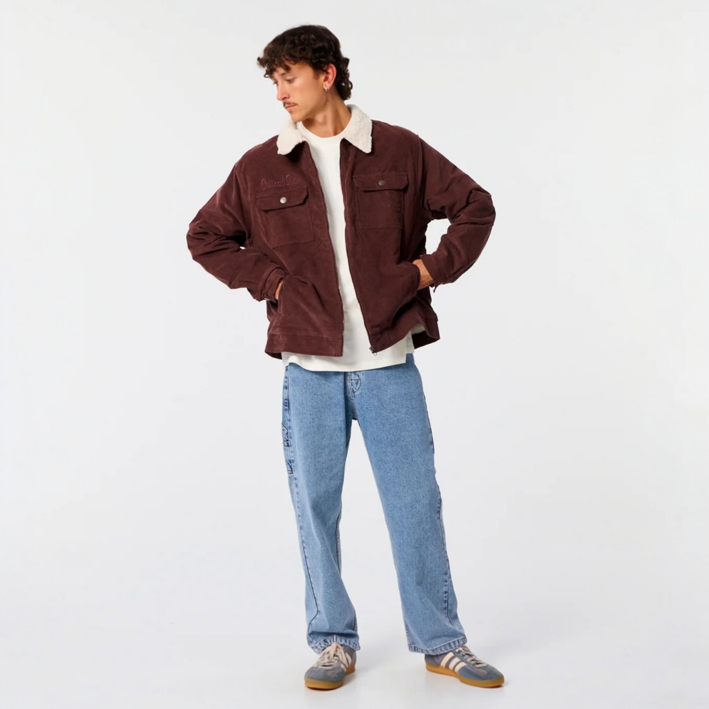 Person wearing a brown jacket with white lining, white shirt, blue jeans, and gray shoes on a white background