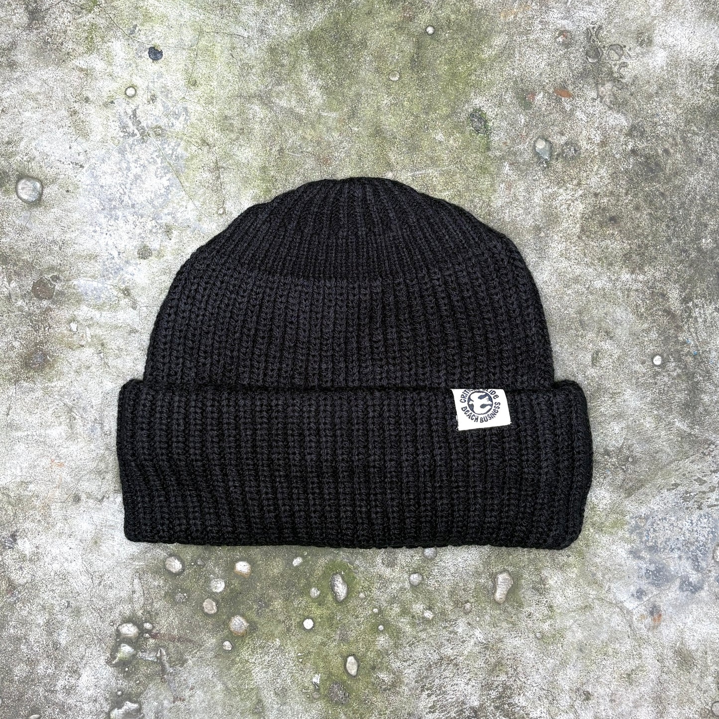 A black ribbed knit beanie hat with a small white embroidered logo patch on the side, laid flat on a textured gray concrete surface.