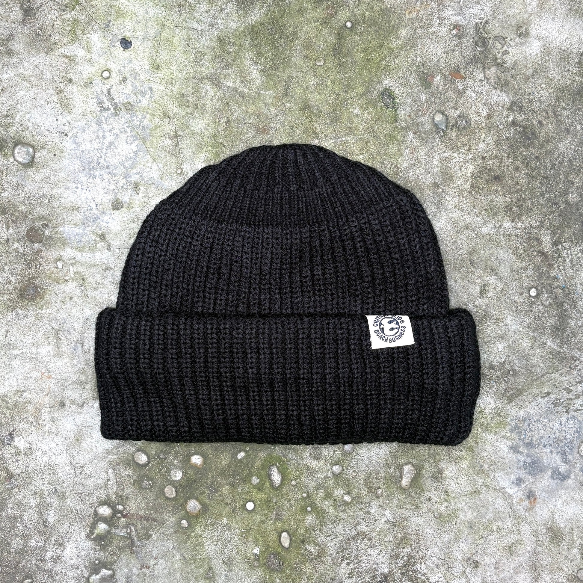 A black ribbed knit beanie hat with a small white embroidered logo patch on the side, laid flat on a textured gray concrete surface.
