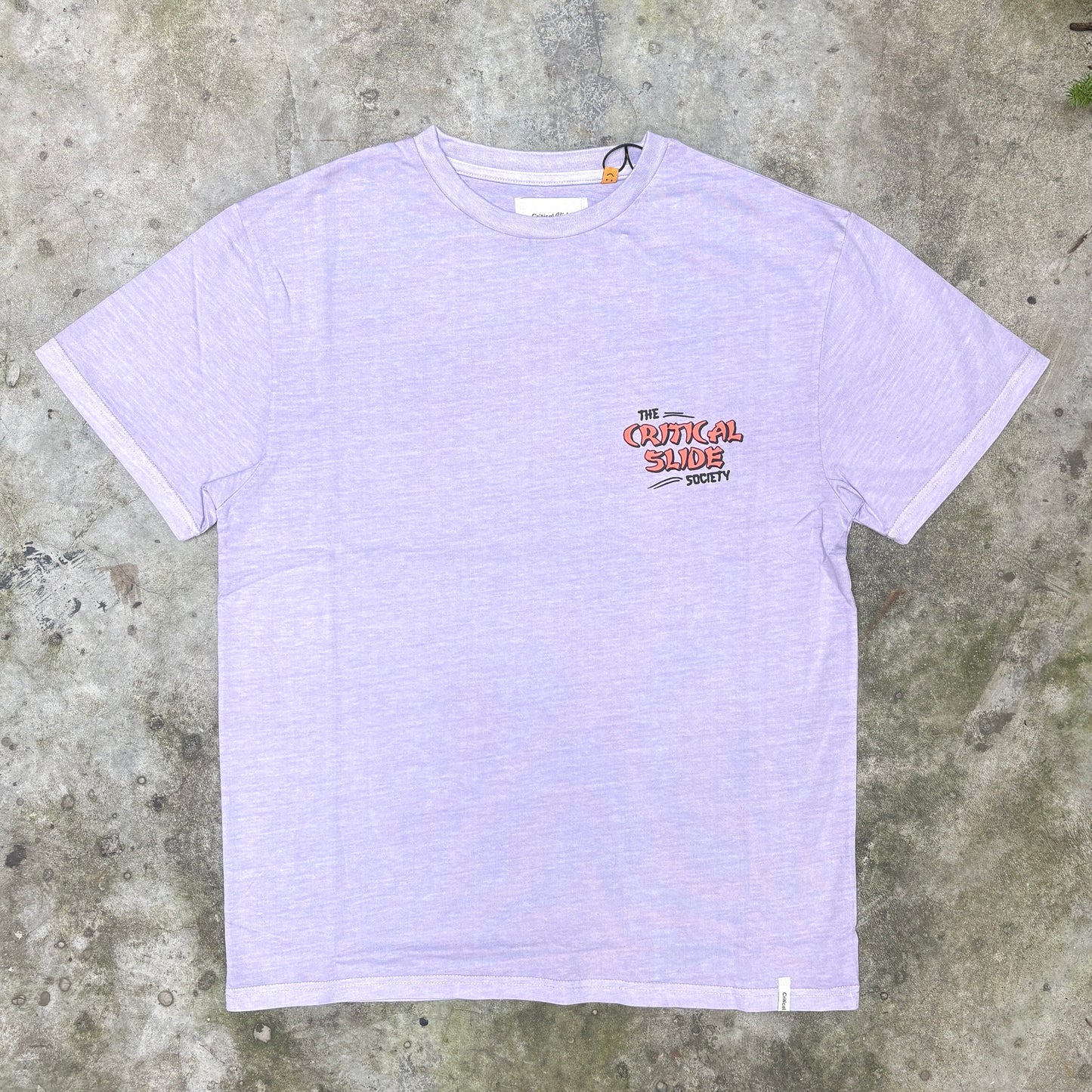 A light lavender heather short-sleeve t-shirt with a small graphic on the left chest reading 'THE CRITICAL SLIDE SOCIETY' in red and black, laid flat on a textured gray concrete surface.