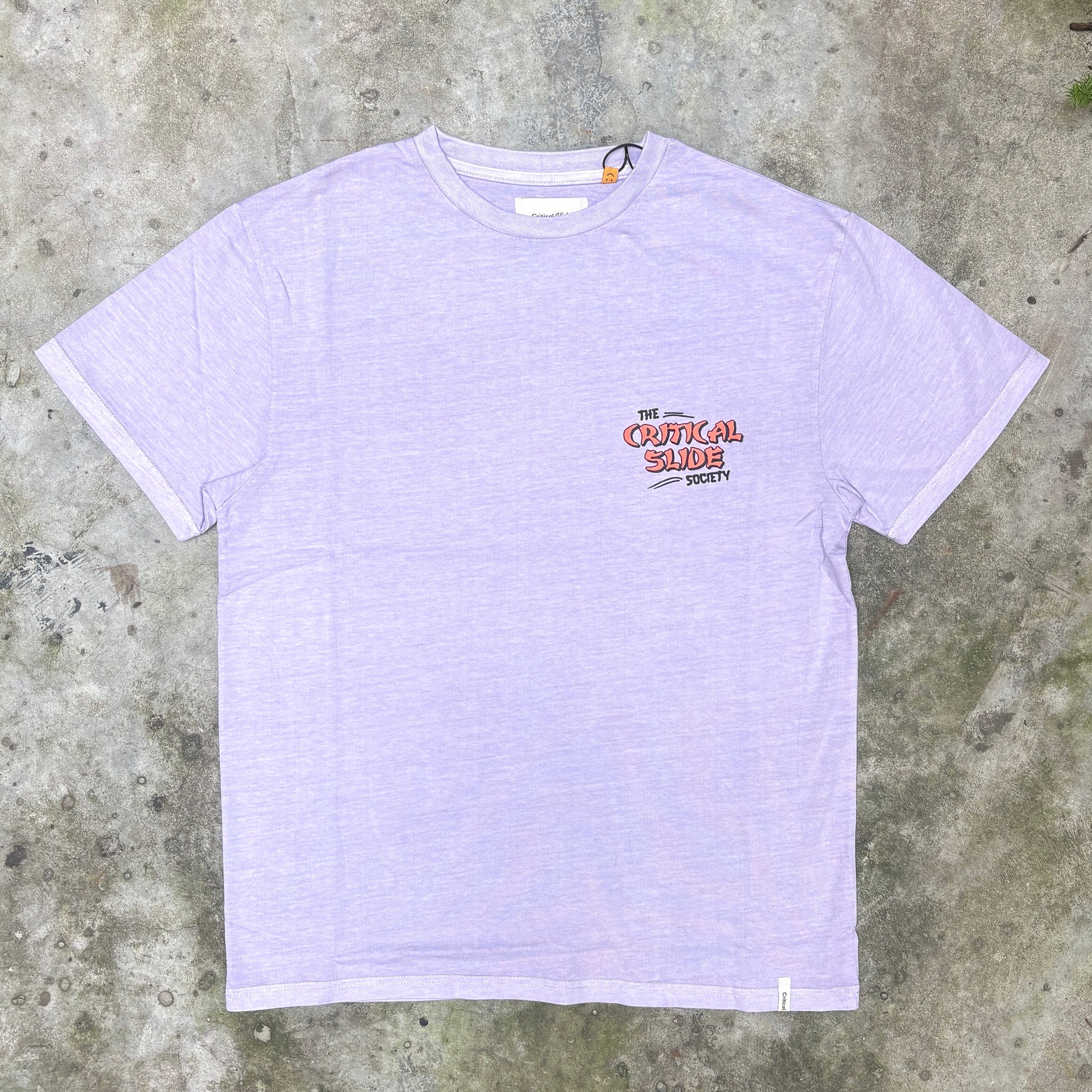 A light lavender heather short-sleeve t-shirt with a small graphic on the left chest reading 'THE CRITICAL SLIDE SOCIETY' in red and black, laid flat on a textured gray concrete surface.
