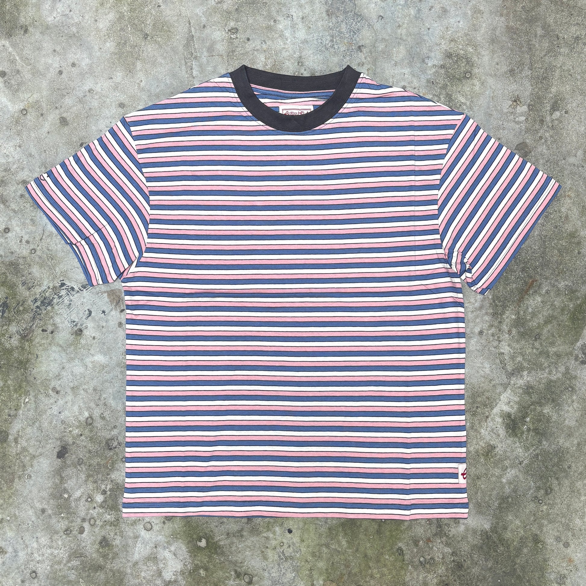 A short-sleeve crewneck t-shirt with horizontal stripes in navy blue, white, and light pink, featuring a solid navy blue ribbed collar, laid flat on a textured gray concrete surface.
