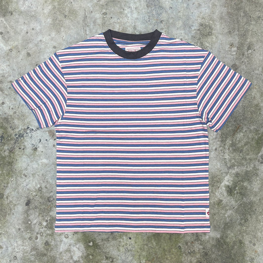 A short-sleeve crewneck t-shirt with horizontal stripes in navy blue, white, and light pink, featuring a solid navy blue ribbed collar, laid flat on a textured gray concrete surface.