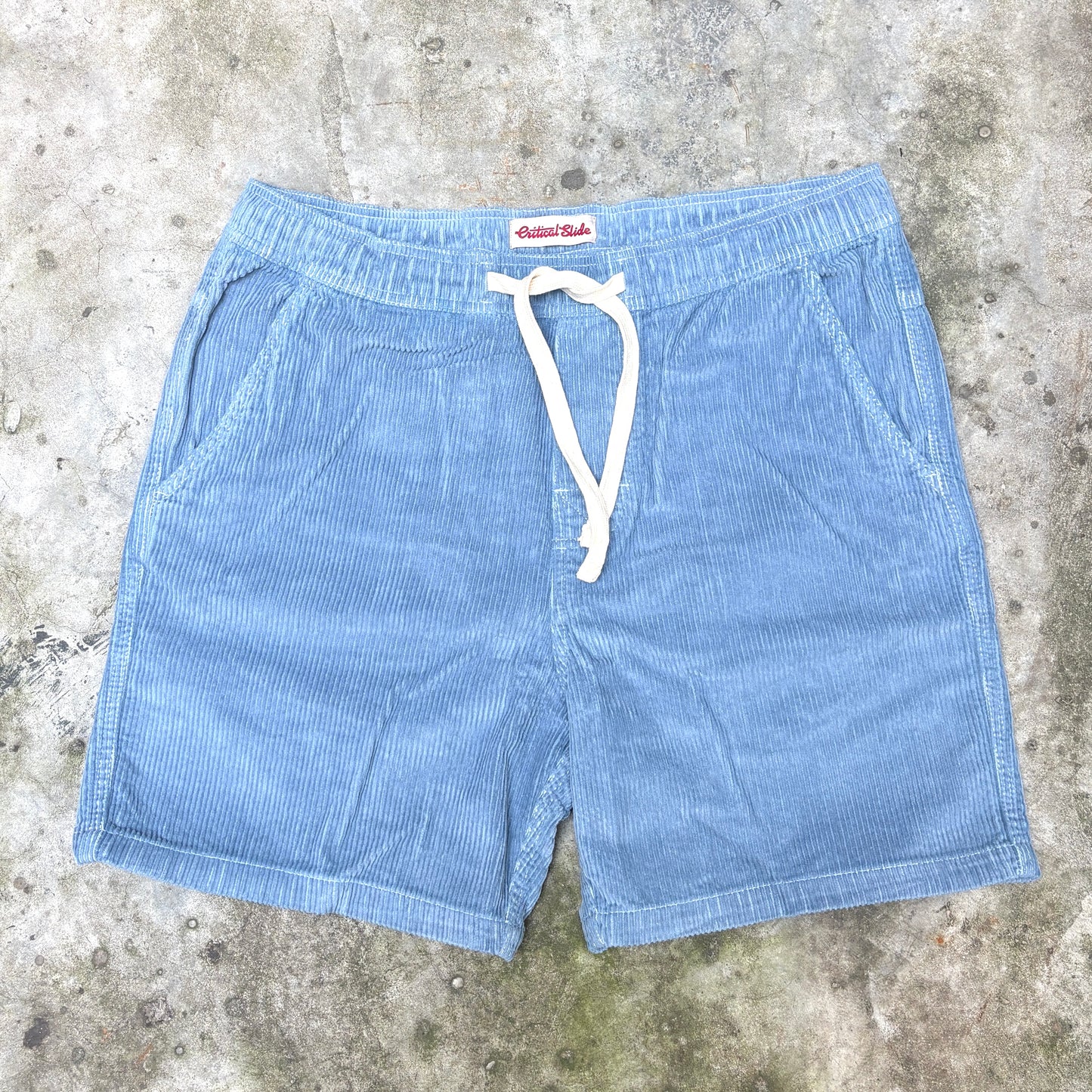 Light blue corduroy shorts with a white drawstring, laid flat on a textured concrete surface. The brand name 'Artist Shot' is visible on the inner waistband label.