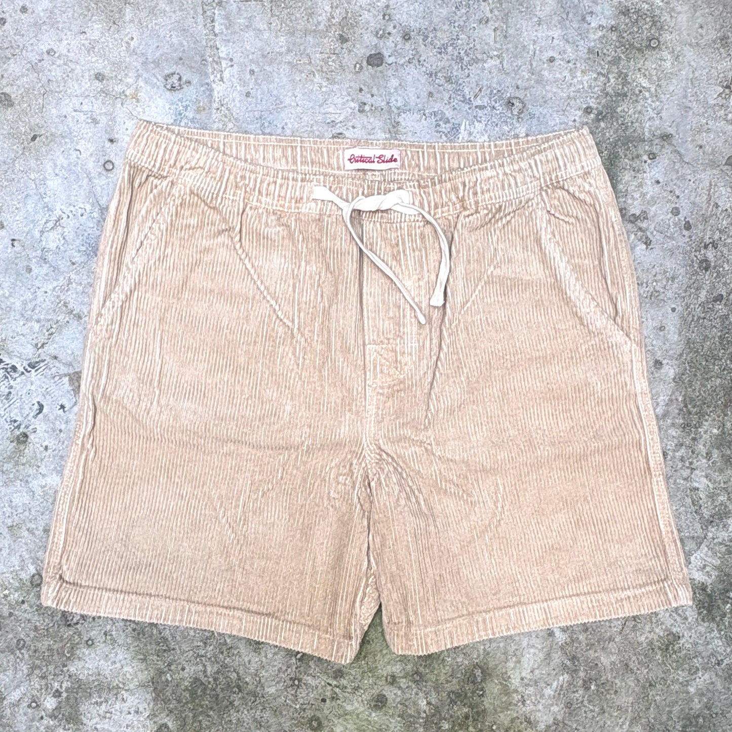 A pair of light beige corduroy shorts with a white drawstring, laid flat on a textured gray concrete surface. The brand label 'Bloomer Club' is visible at the waistband.