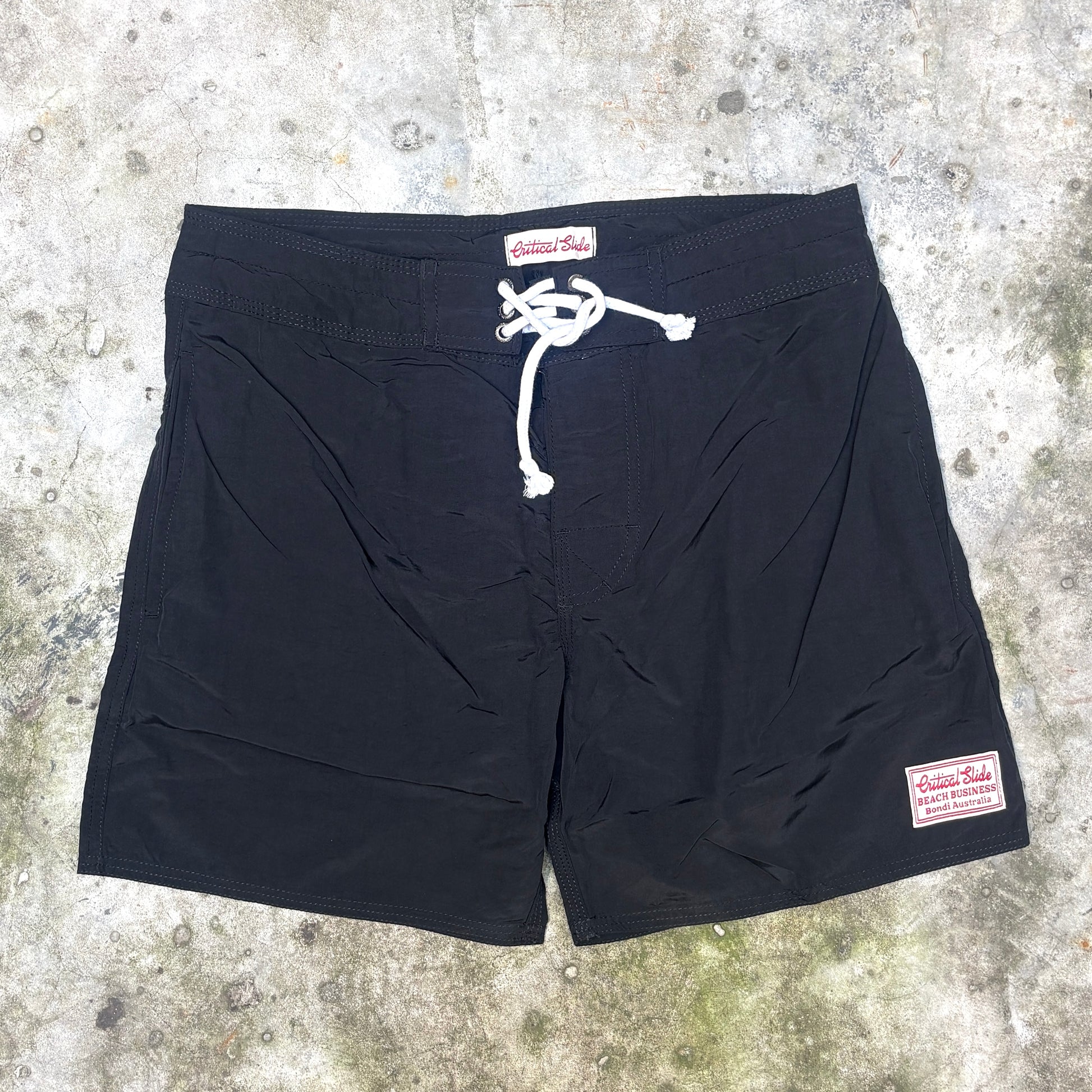Black board shorts with a white drawstring and a visible 'Historic Roads' brand patch, laid flat on a textured concrete surface.