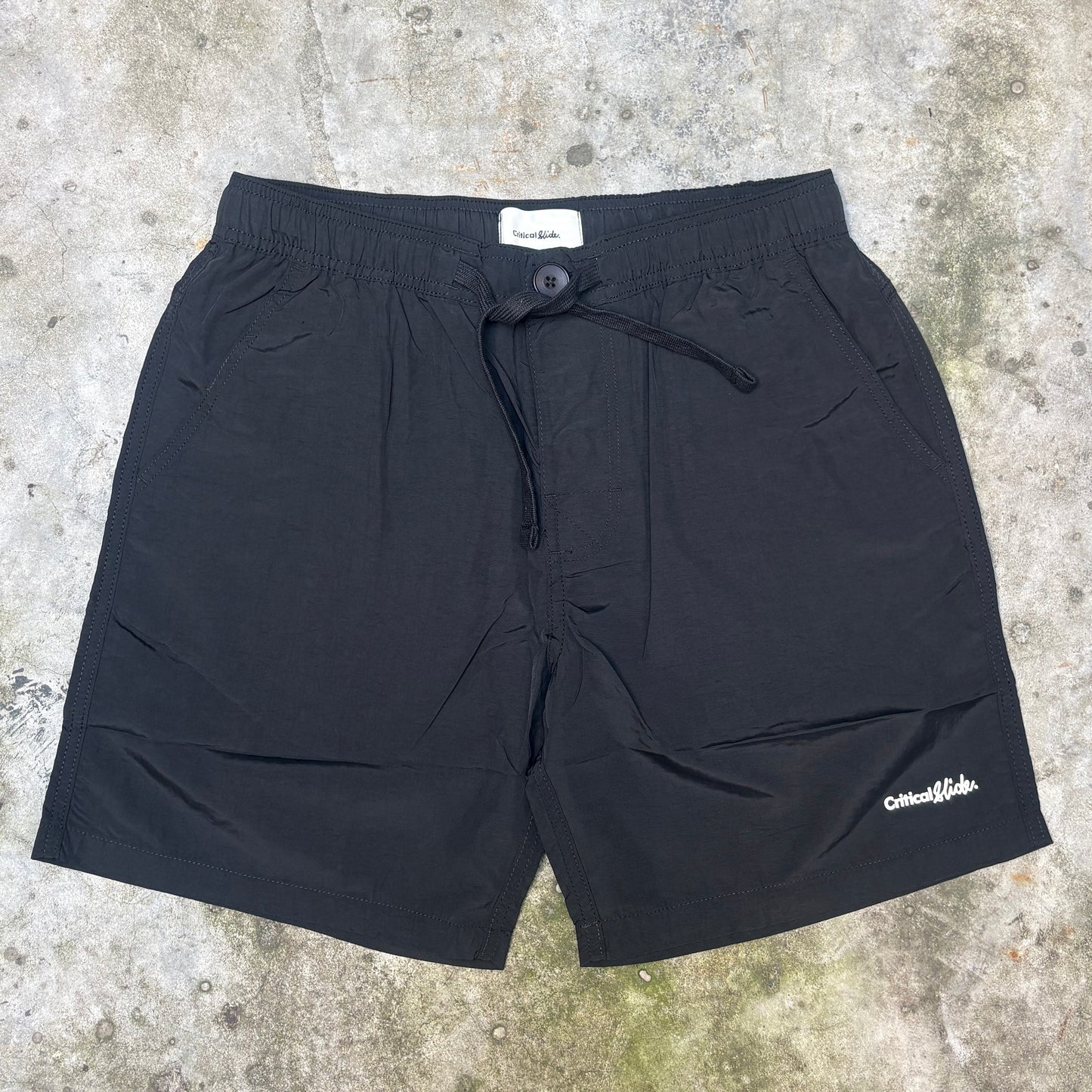 Black drawstring shorts laid flat on a textured concrete surface, with a small white script logo near the lower right leg.