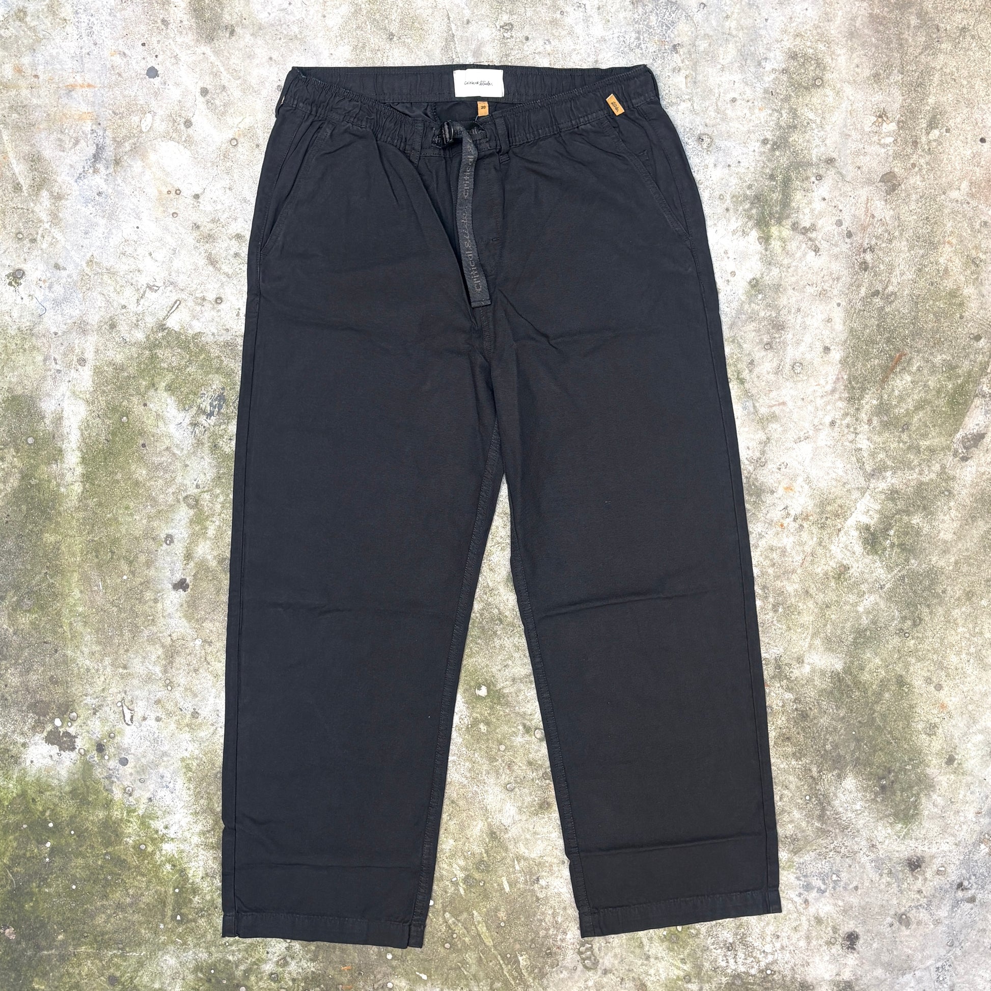 Black wide-leg cropped pants with an elastic drawstring waist, laid flat on a textured concrete surface.