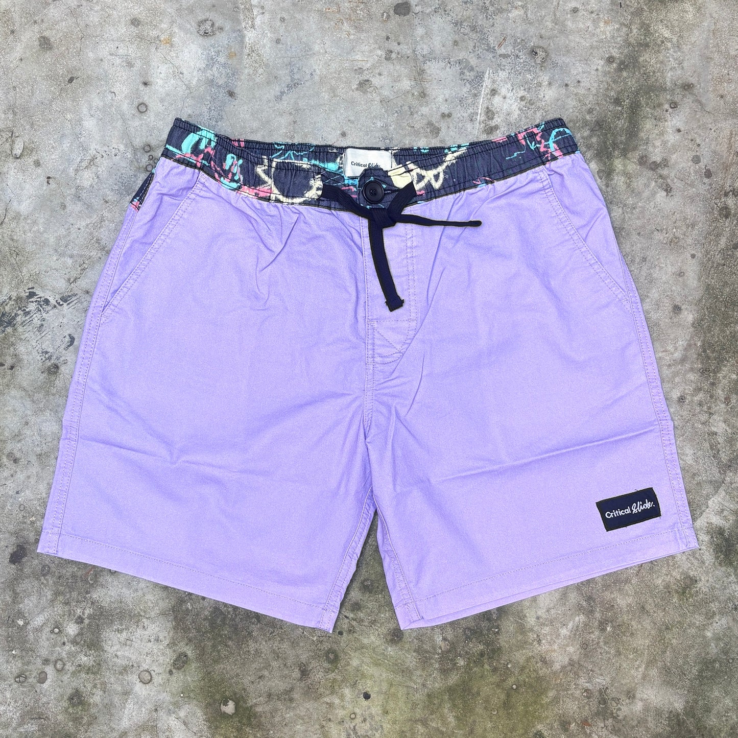 Purple board shorts with a multicolor patterned elastic waistband and black drawstring, laid flat on a gray concrete surface. A black 'Columbia' brand patch is visible on the lower right leg.
