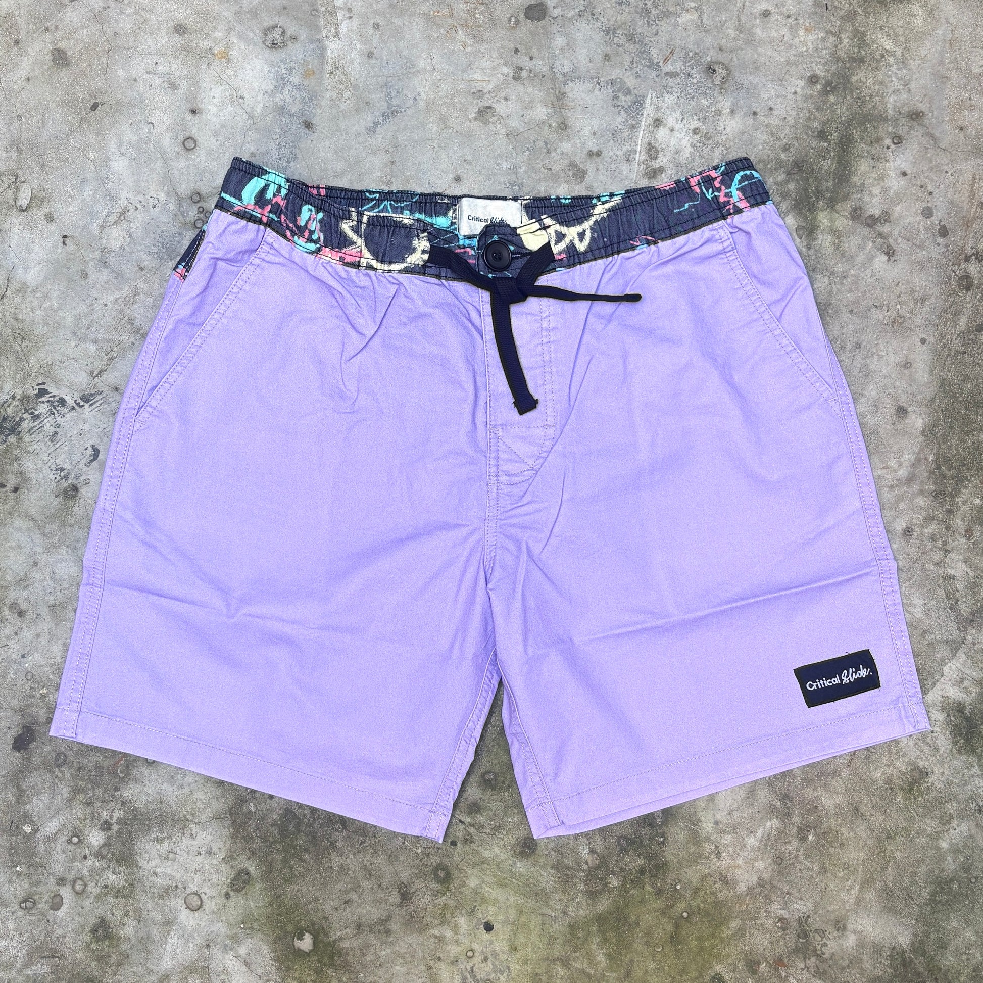 Purple board shorts with a multicolor patterned elastic waistband and black drawstring, laid flat on a gray concrete surface. A black 'Columbia' brand patch is visible on the lower right leg.
