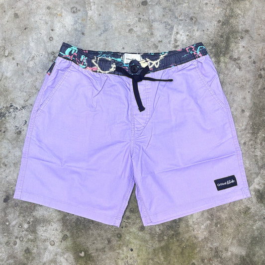 Purple board shorts with a multicolor patterned elastic waistband and black drawstring, laid flat on a gray concrete surface. A black 'Columbia' brand patch is visible on the lower right leg.