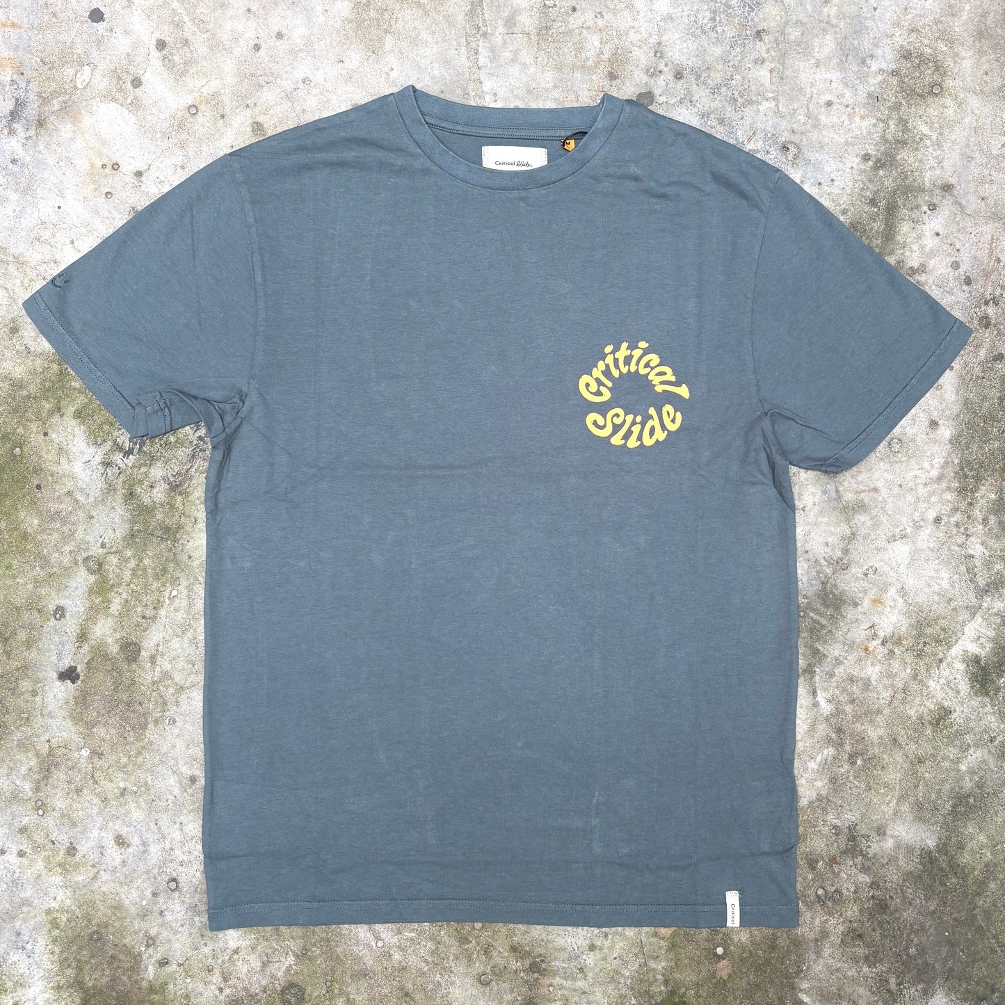 A slate blue crewneck t-shirt with a yellow 'Critical Guide' logo printed on the left chest, laid flat on a textured concrete surface.
