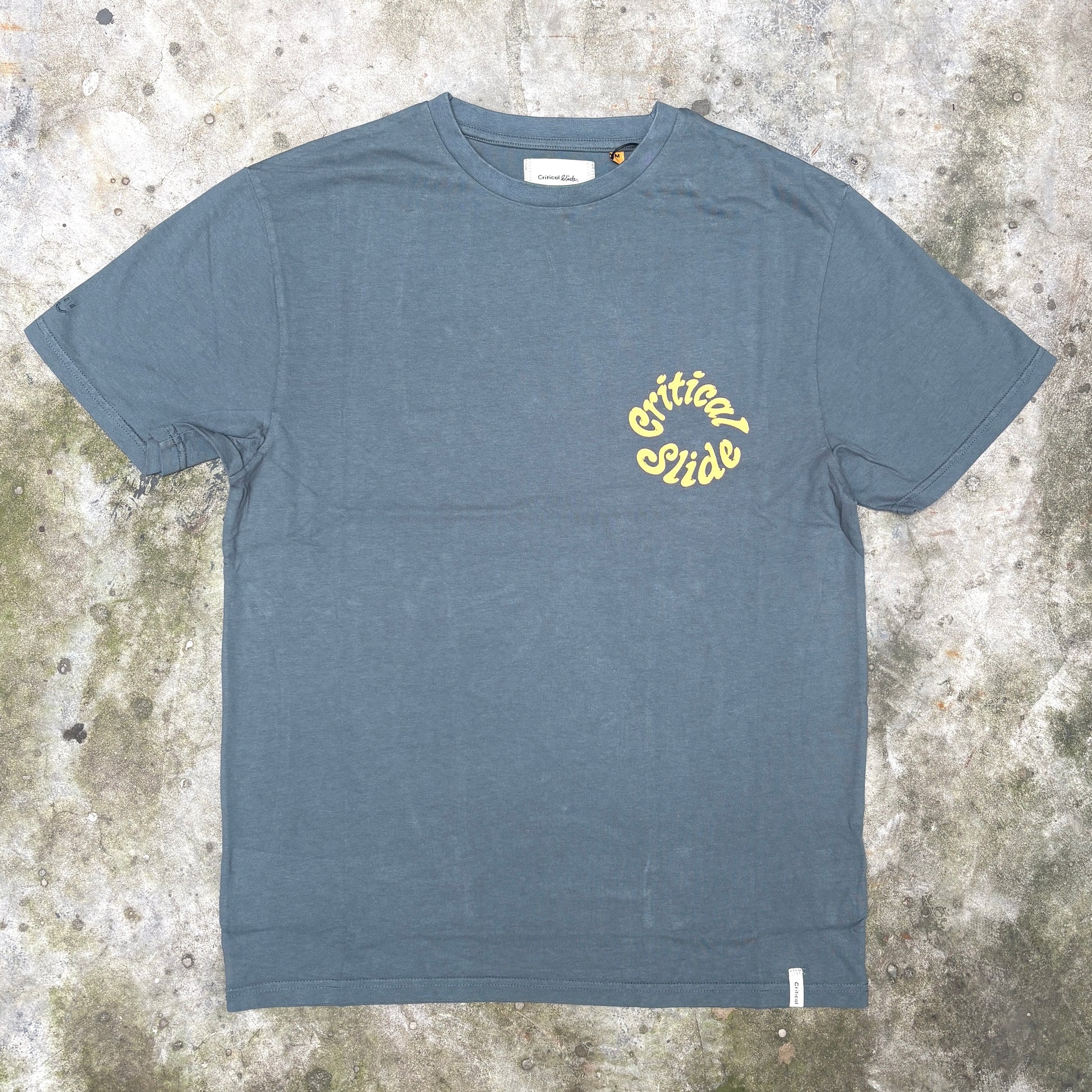 A slate blue crewneck t-shirt with a yellow 'Critical Guide' logo printed on the left chest, laid flat on a textured concrete surface.