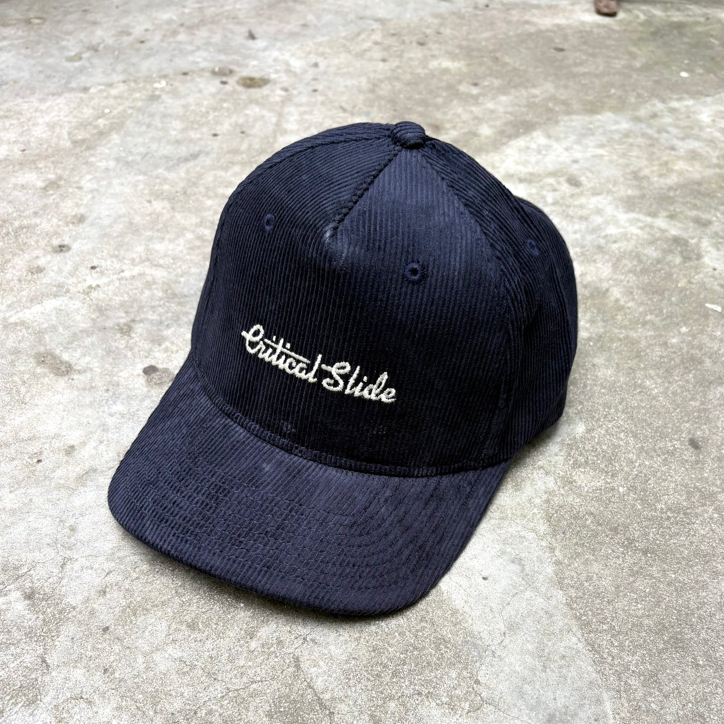 Blue cap with 'Critical Slide' embroidery on a concrete surface