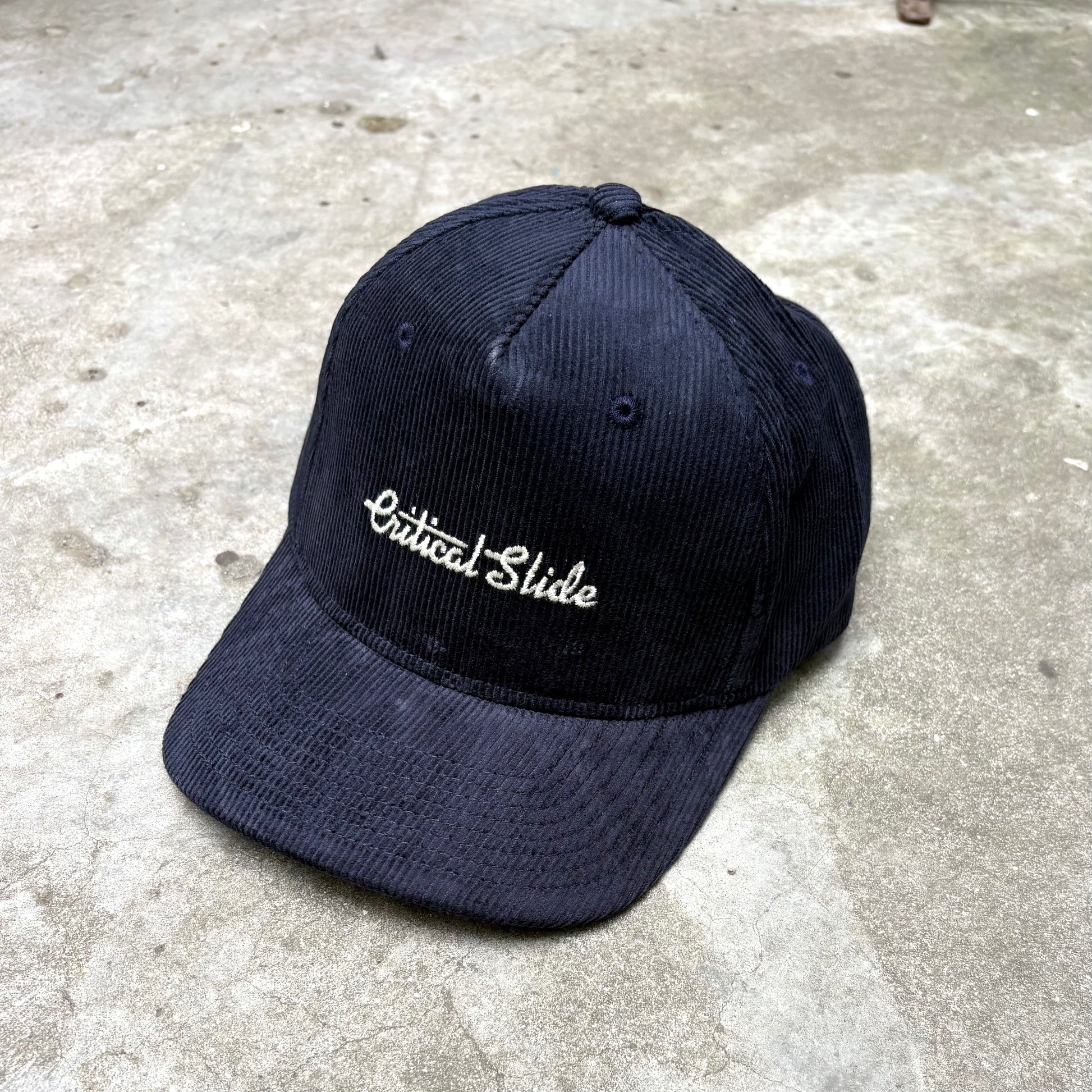 Blue cap with 'Critical Slide' embroidery on a concrete surface